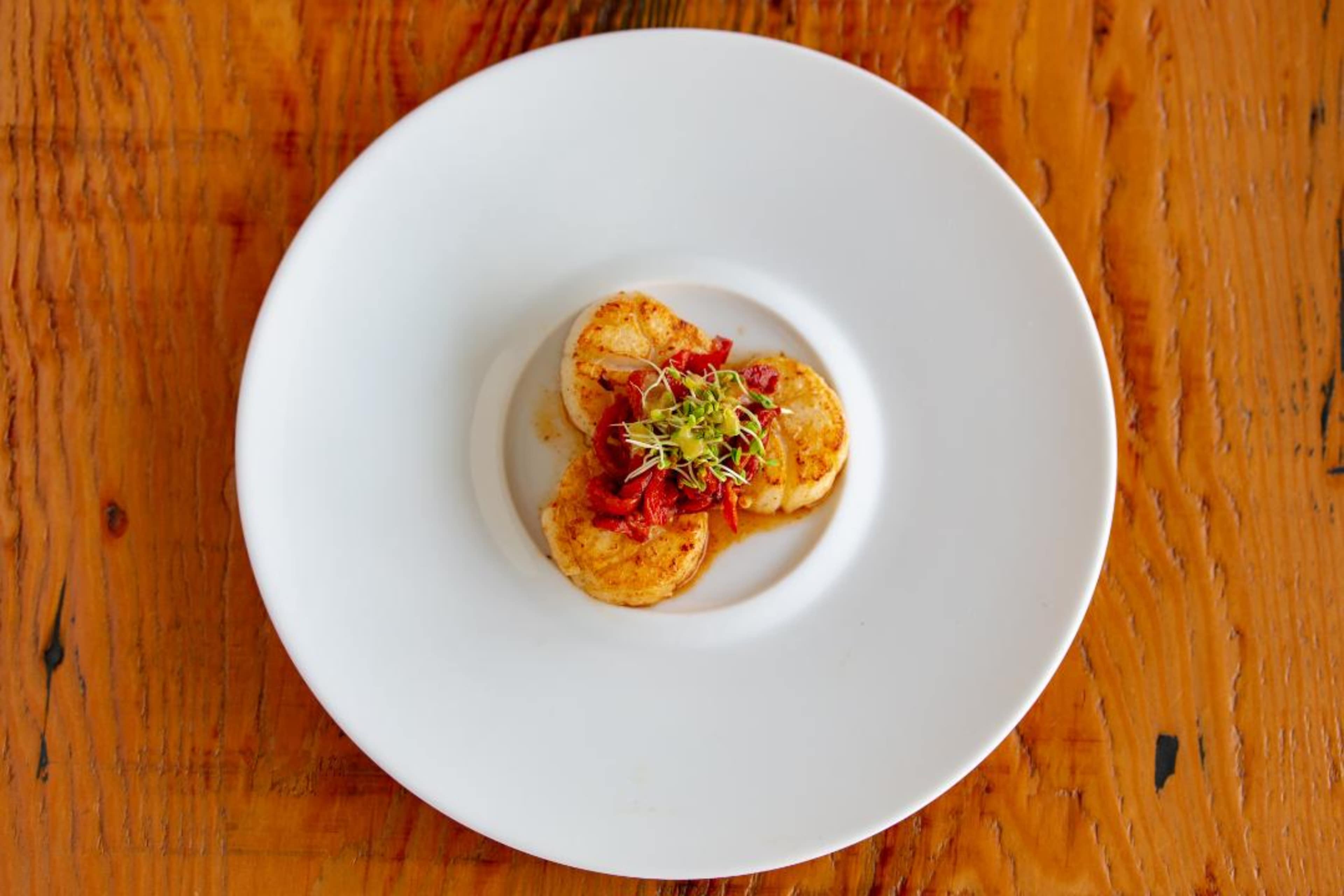 A plate with three round pieces of fried food topped with red sauce and garnished with sprouts rests on a wooden table.