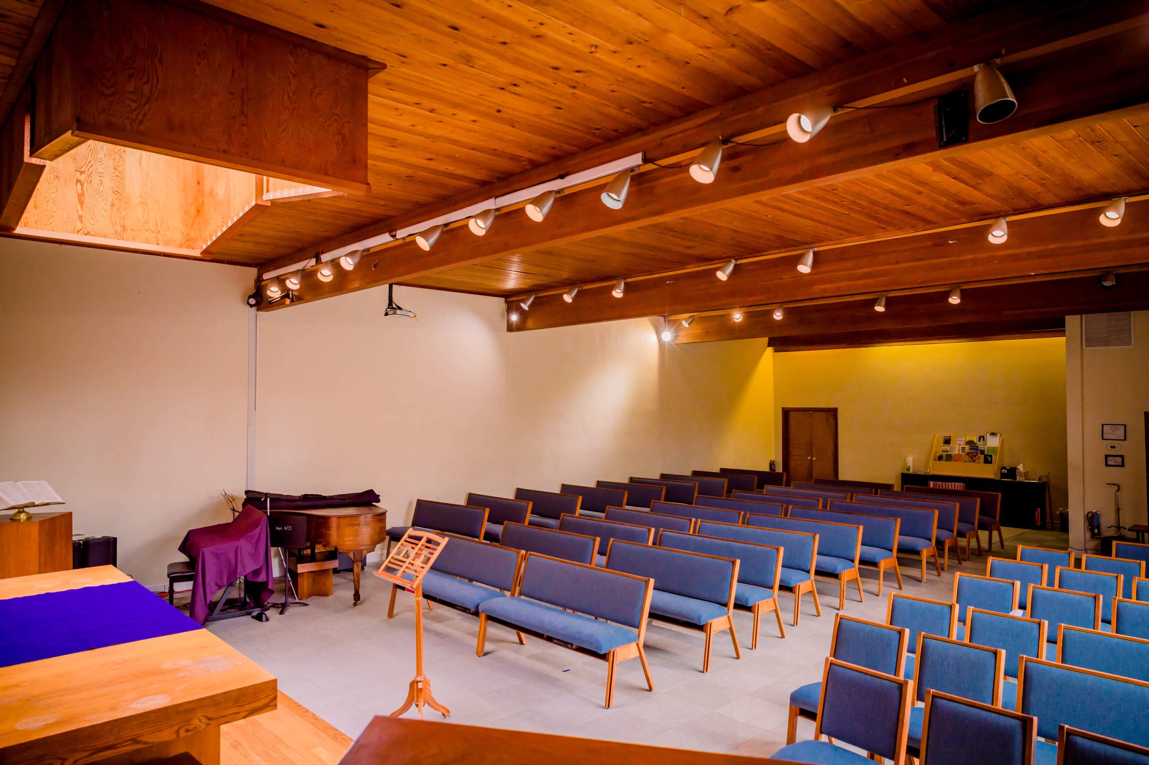 The image depicts an interior of a simple auditorium with blue upholstered seating arranged in rows beneath wooden beams and ceiling lights.