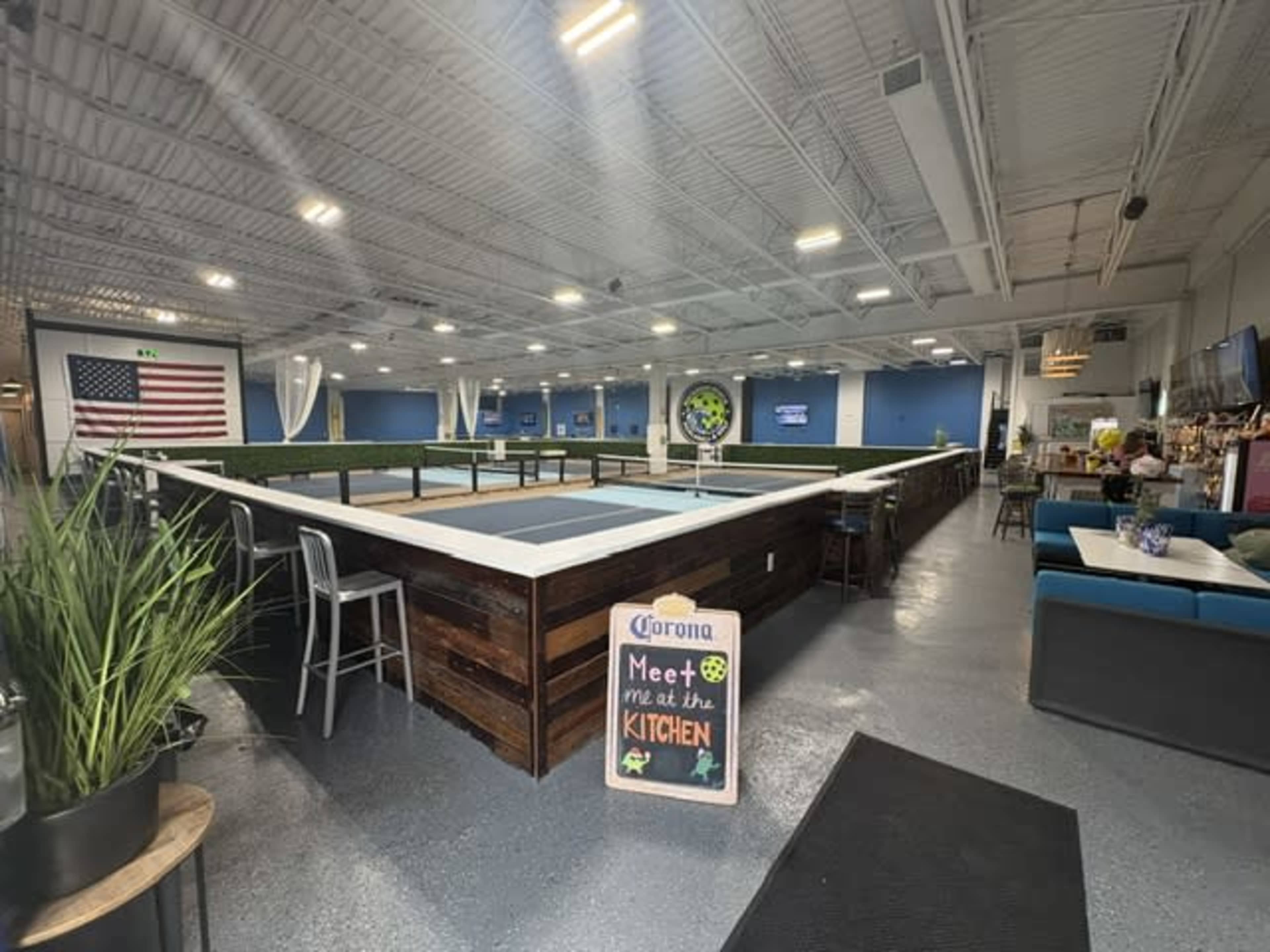 The image shows the interior of a spacious venue featuring a large, rectangular table in the center, surrounded by seating areas and decor, with an American flag displayed on one wall.