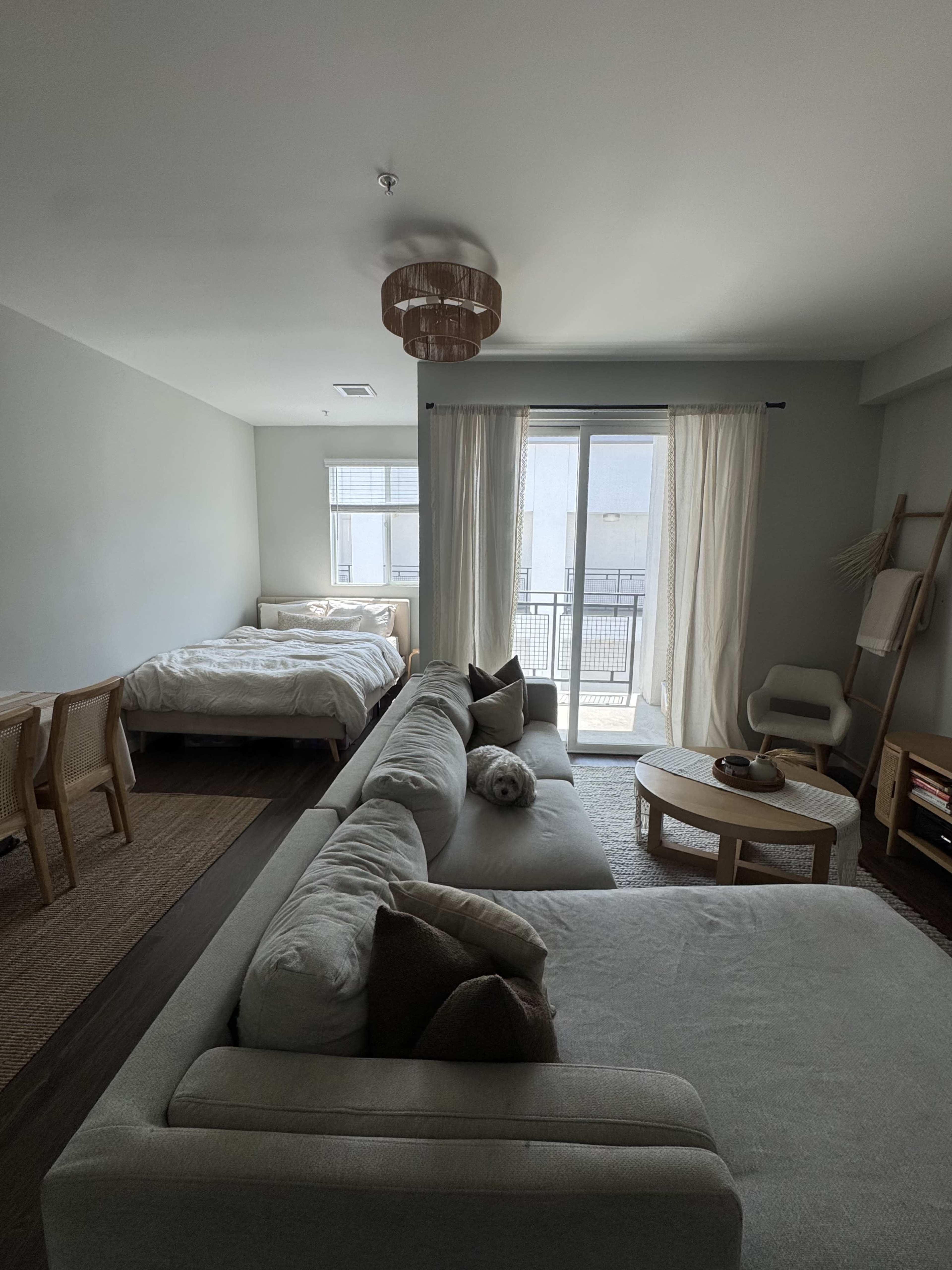 The image shows a modern, open-concept living space with a bed in the corner, a large sectional sofa, a circular coffee table, and a balcony visible through sliding glass doors.