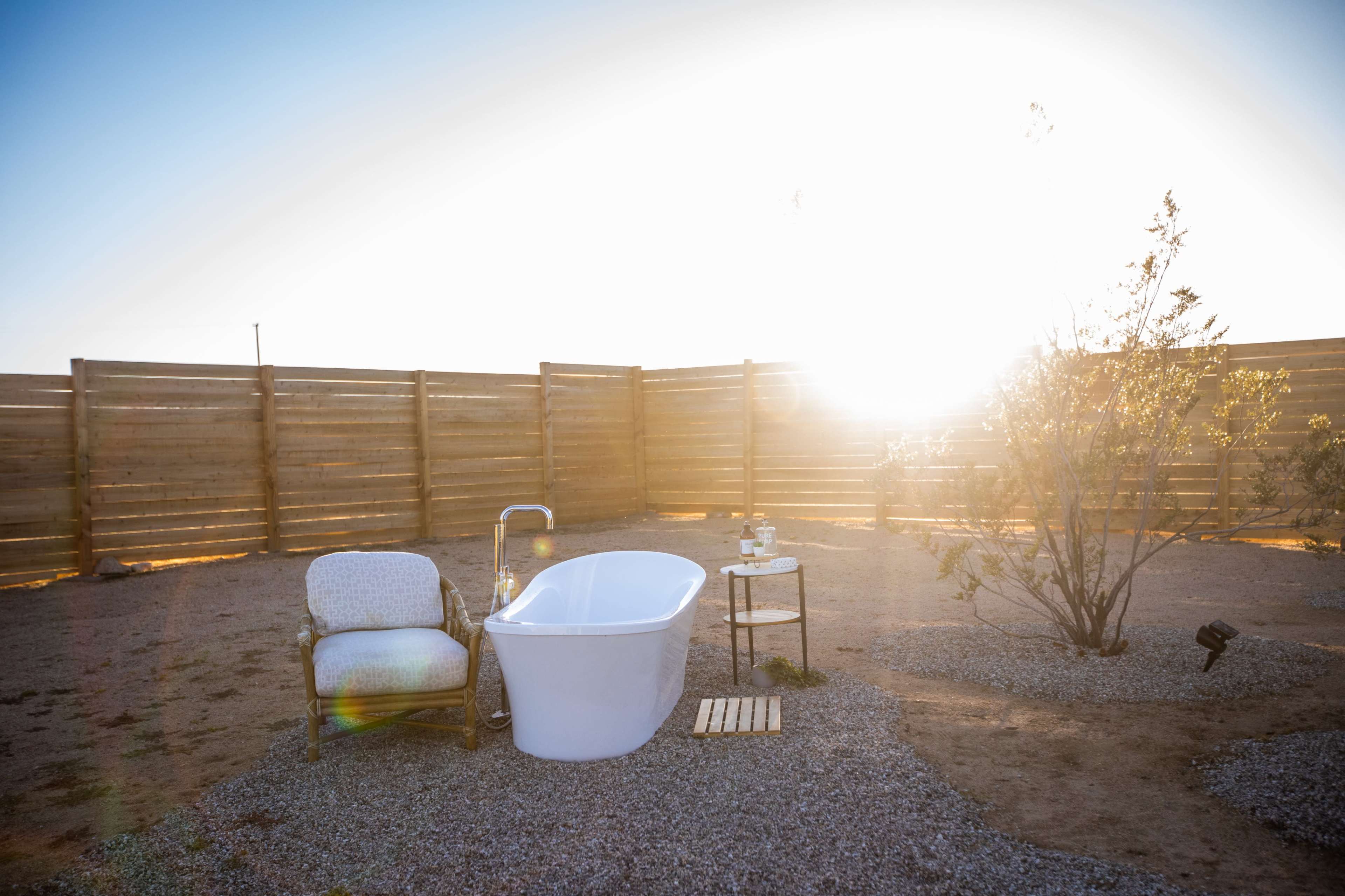 A white freestanding bathtub and a chair are set on a gravel area surrounded by a wooden fence, with the sun shining in the background.