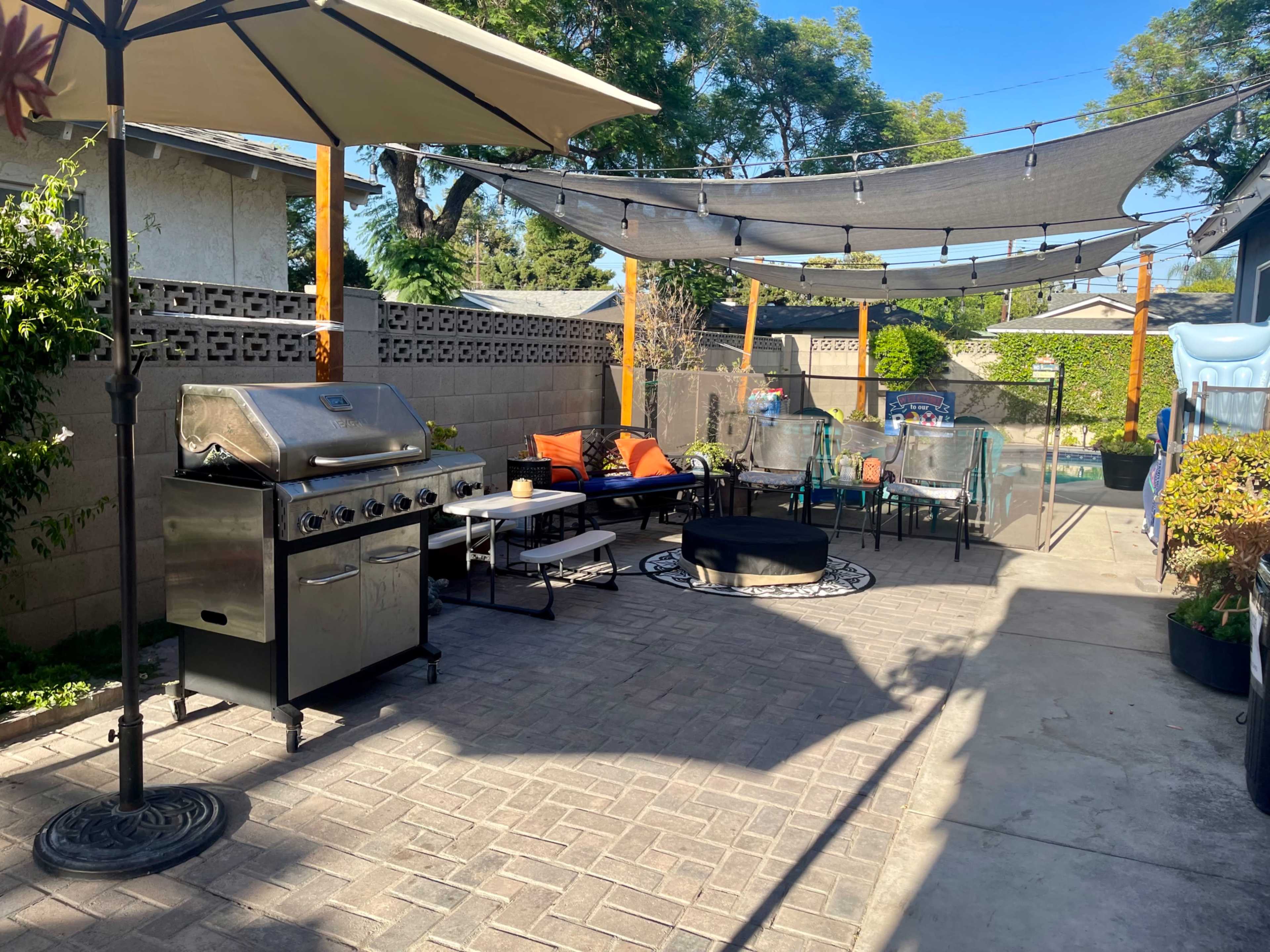 The outdoor patio features a stainless steel grill, comfortable seating, and shaded areas with a fire pit and a swimming pool visible in the background.