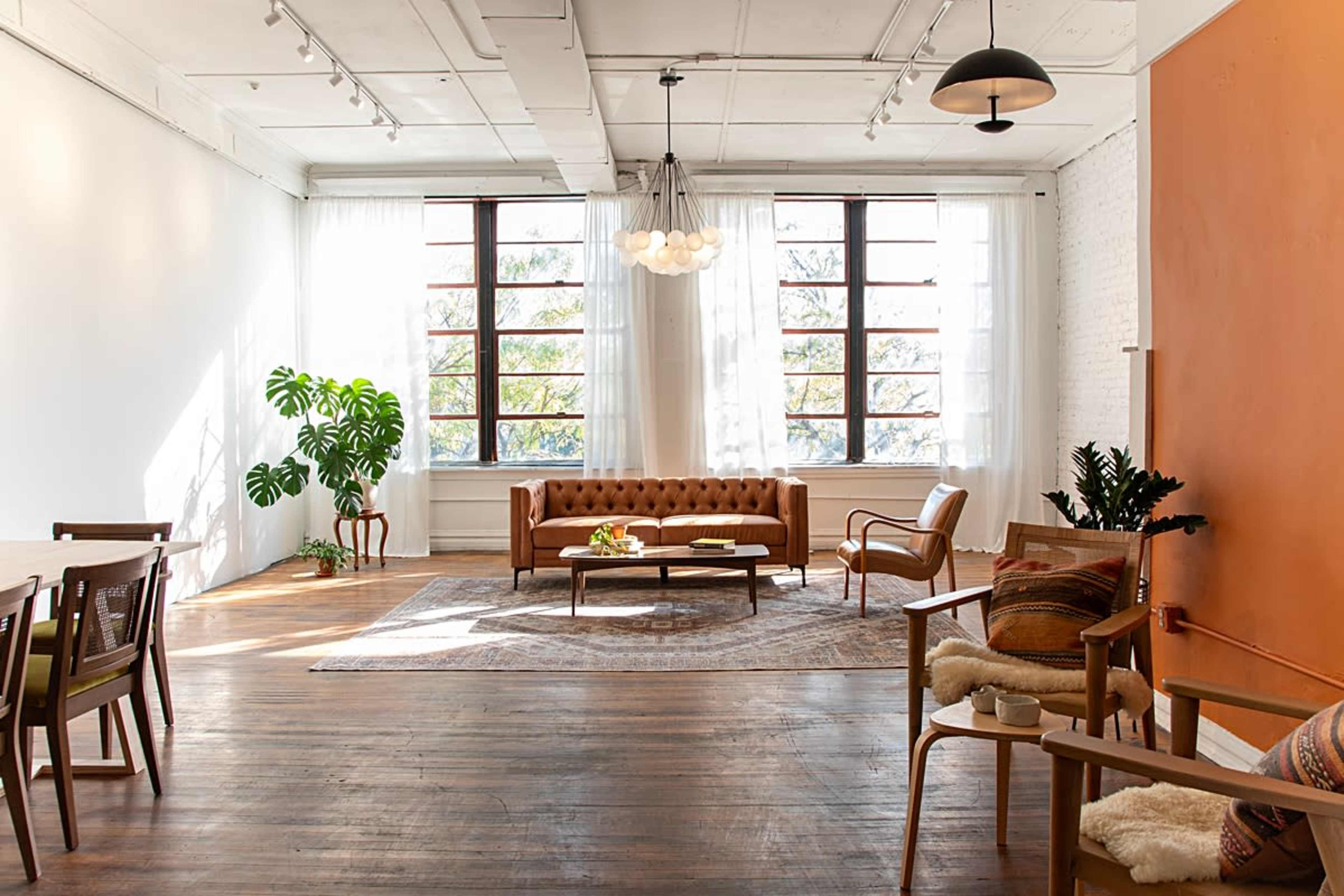The image shows a modern living space with a brown leather sofa, wooden dining furniture, and large windows allowing natural light to enter.