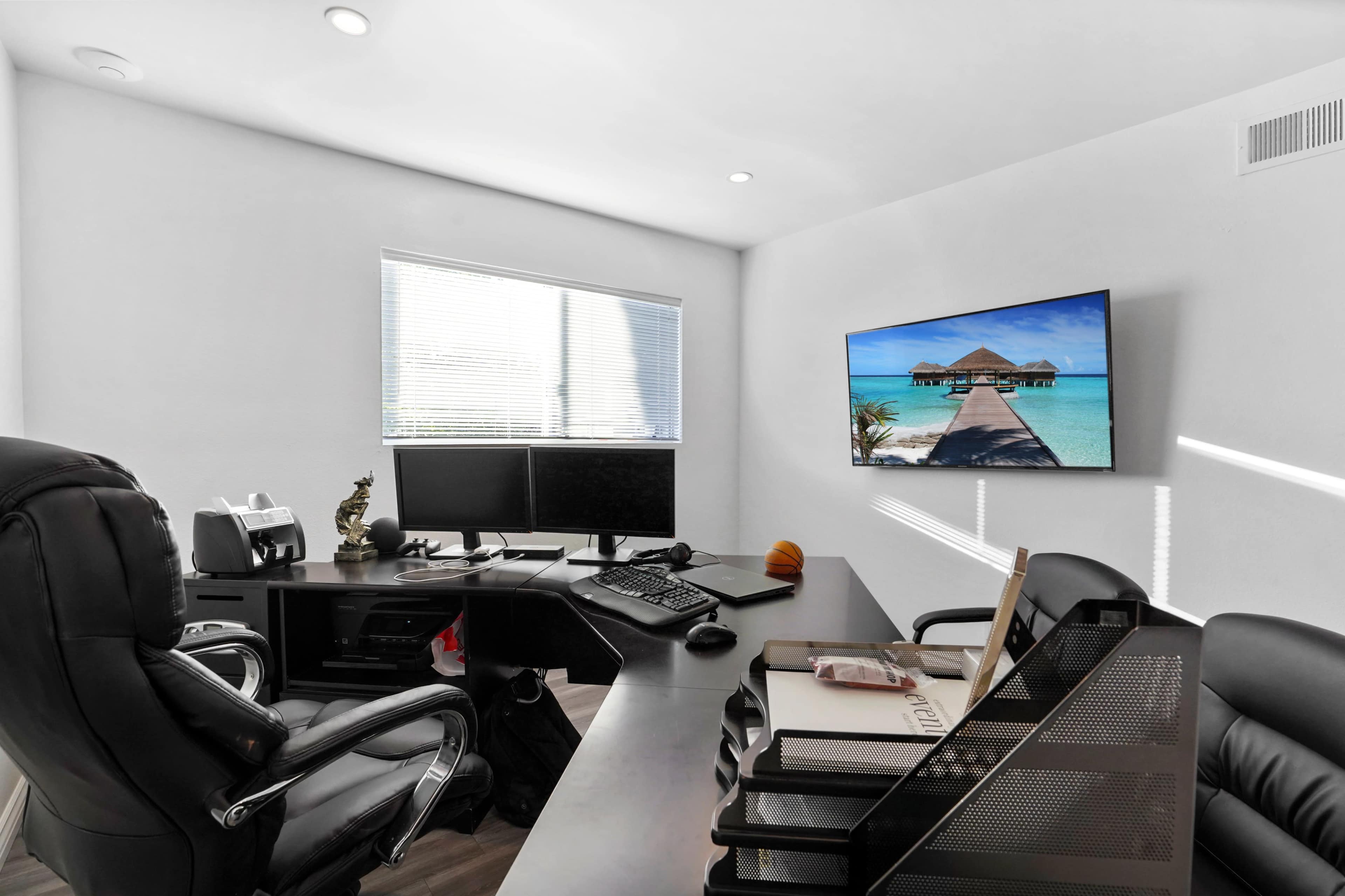 The image shows a modern office space with a black leather chair, two computer monitors on a desk, and a television mounted on the wall displaying a beach scene.