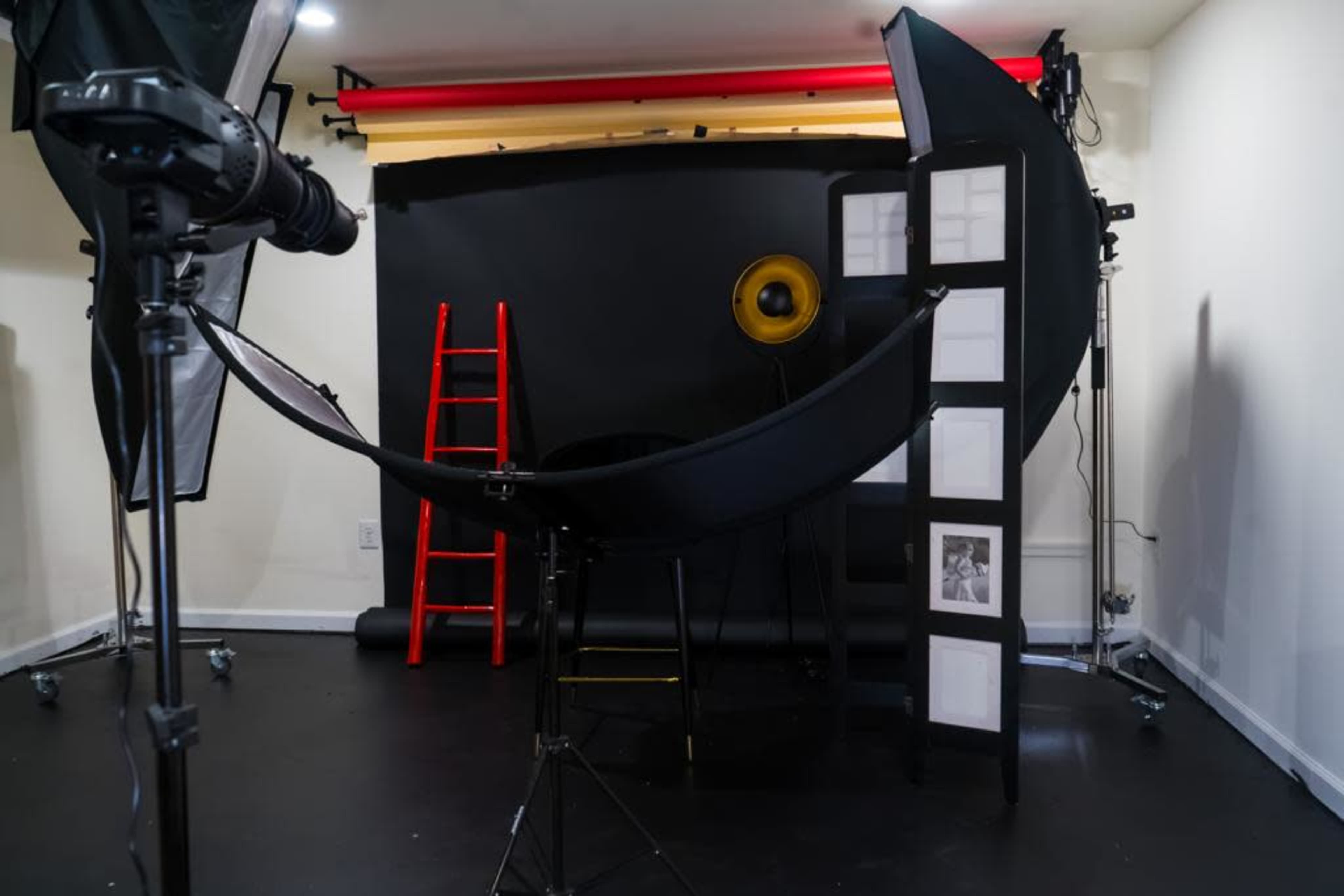 A photography studio setup with various equipment, including a black backdrop, a microphone stand, a ladder, and a large circular reflector.