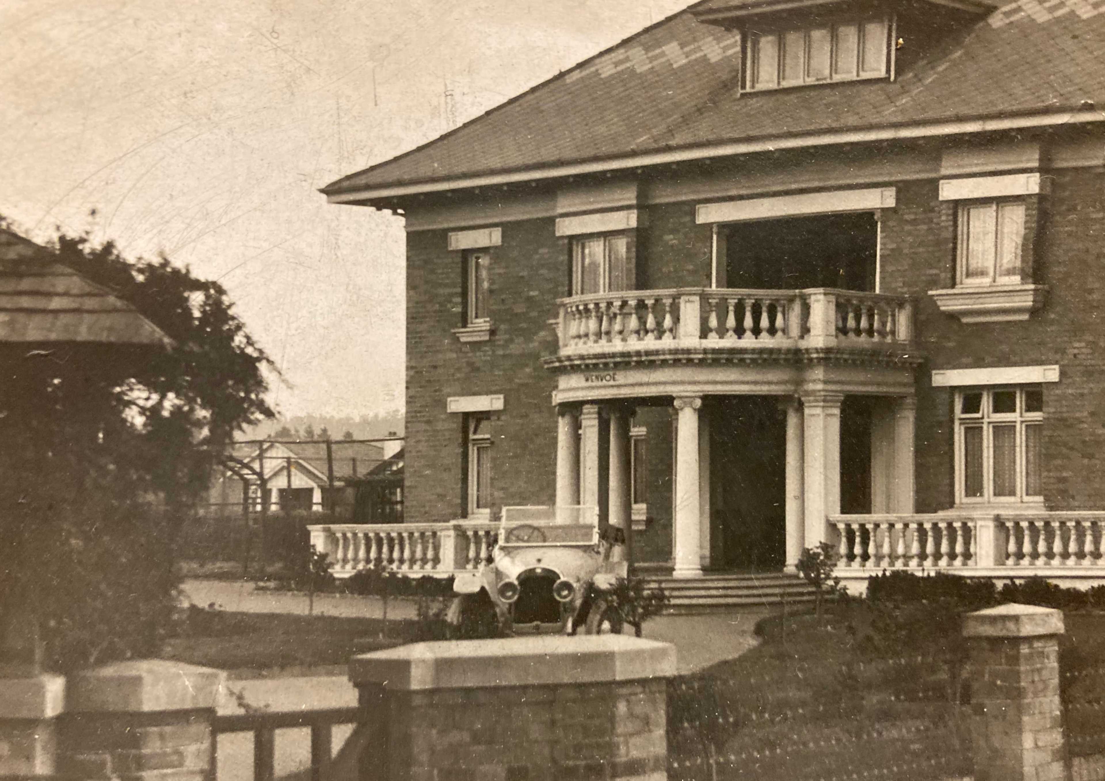 A two-story brick house with a balcony and a vintage car parked in front.