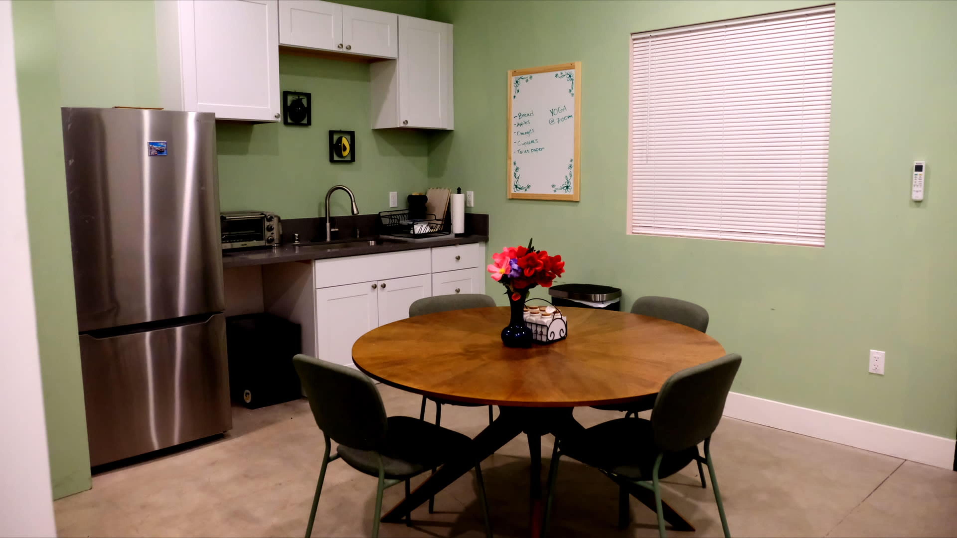 The image shows a kitchen and dining area featuring a round wooden table with four chairs, a metal fridge, and green walls adorned with a whiteboard and a floral centerpiece.