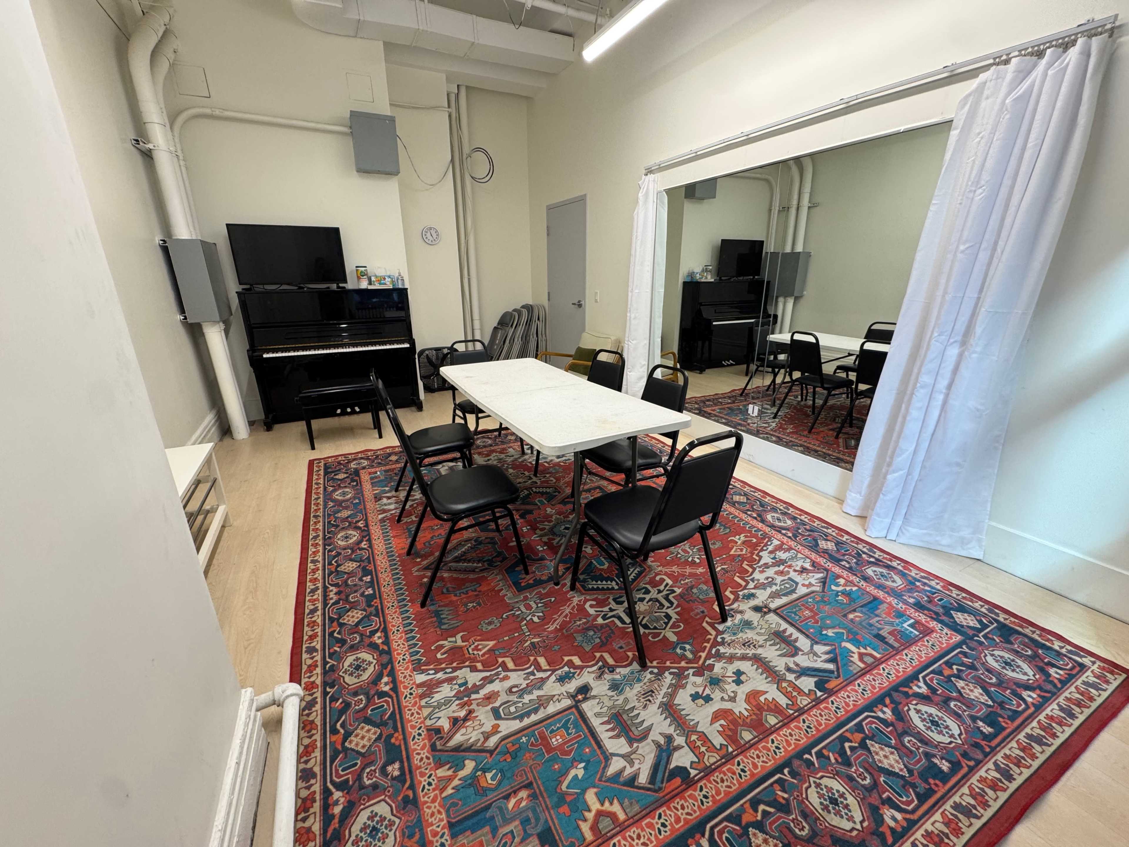 The image shows a room with a large rug, six black chairs arranged around a white table, a piano in the corner, and a mirror along one wall.