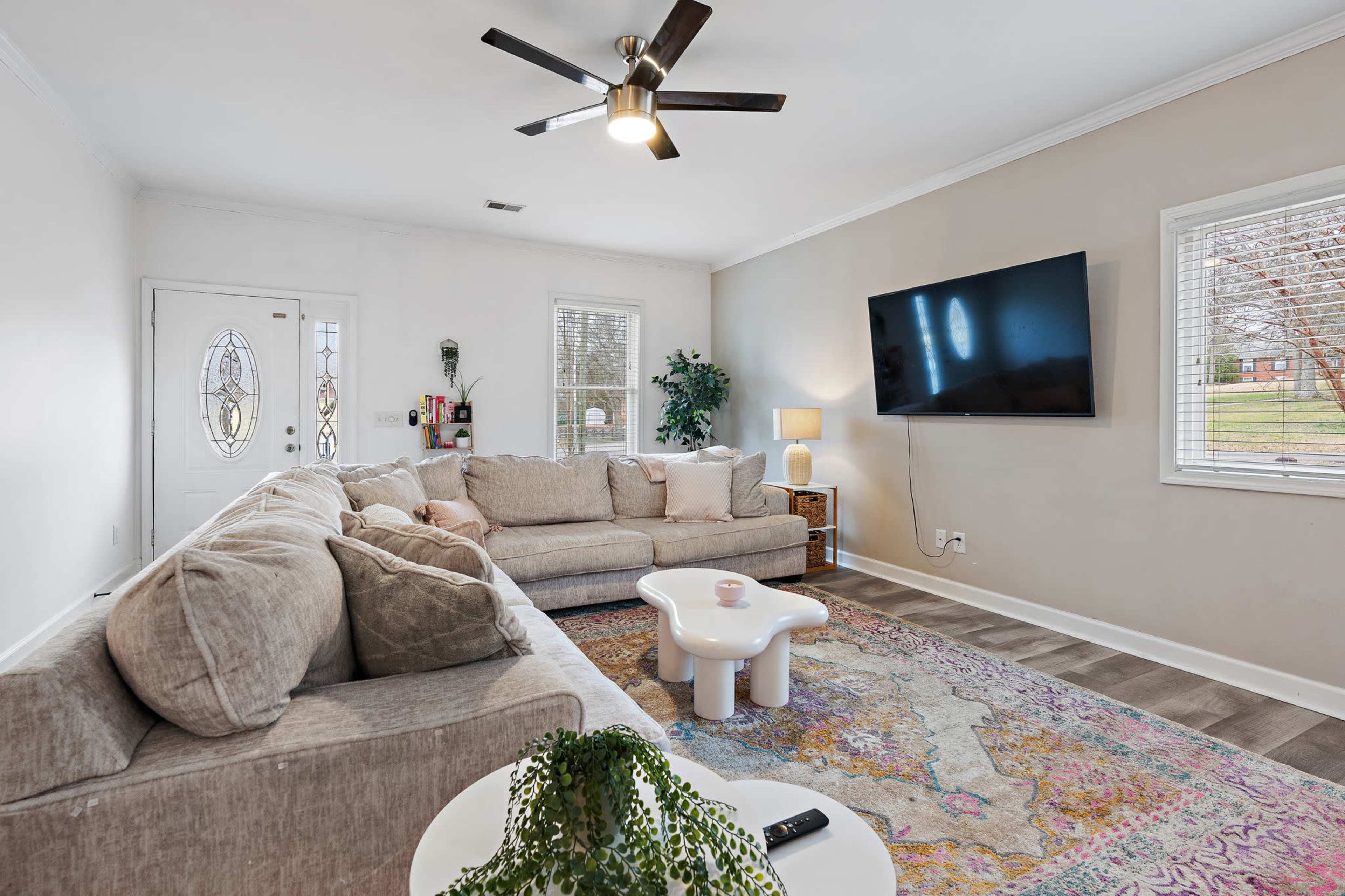 A spacious living room features a large sectional sofa, a round coffee table, a mounted TV, and natural light streaming through the windows.