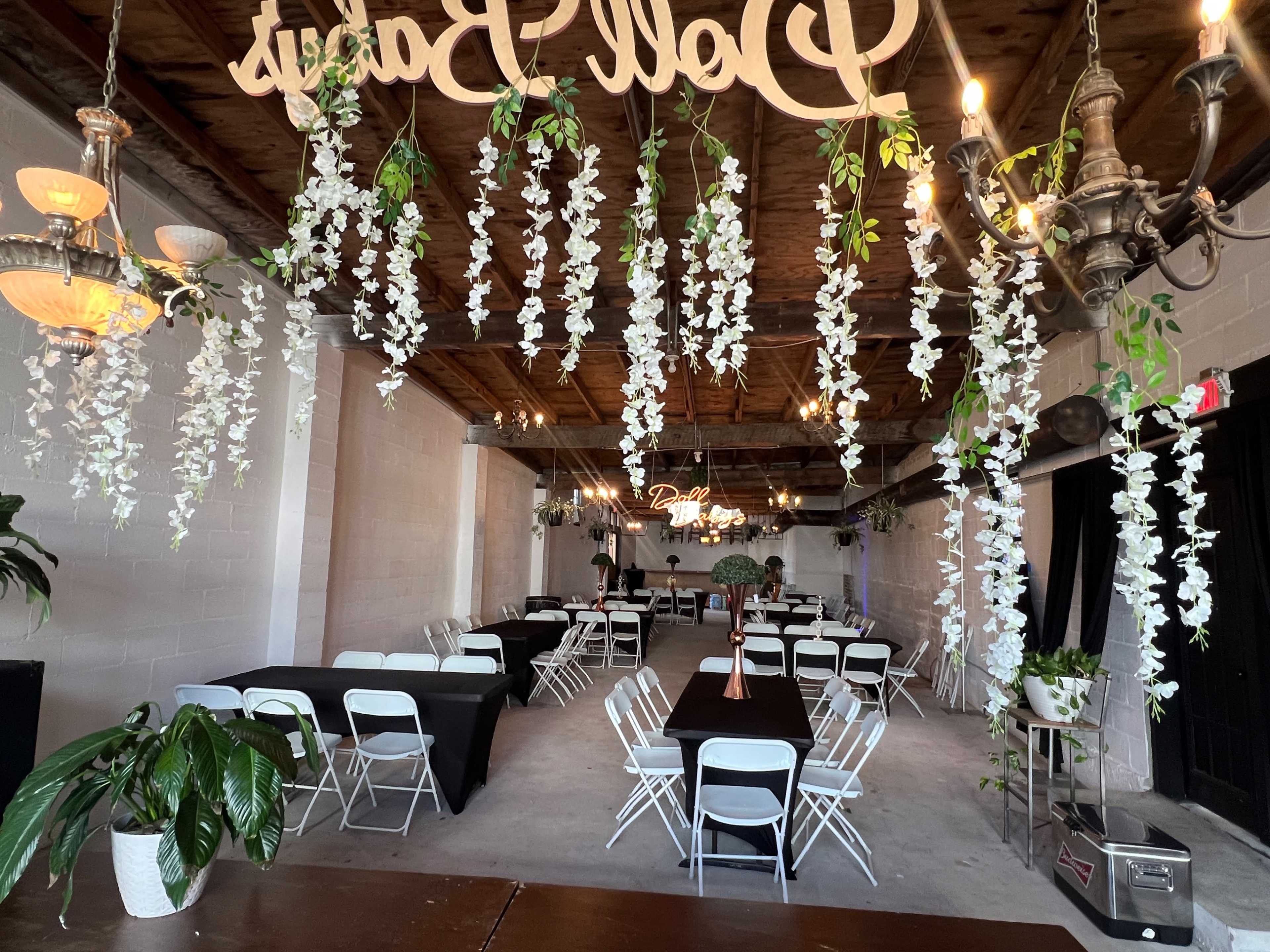 The image shows a spacious event venue with white foldable chairs arranged around black tables, and decorative white flowers hanging from the ceiling.