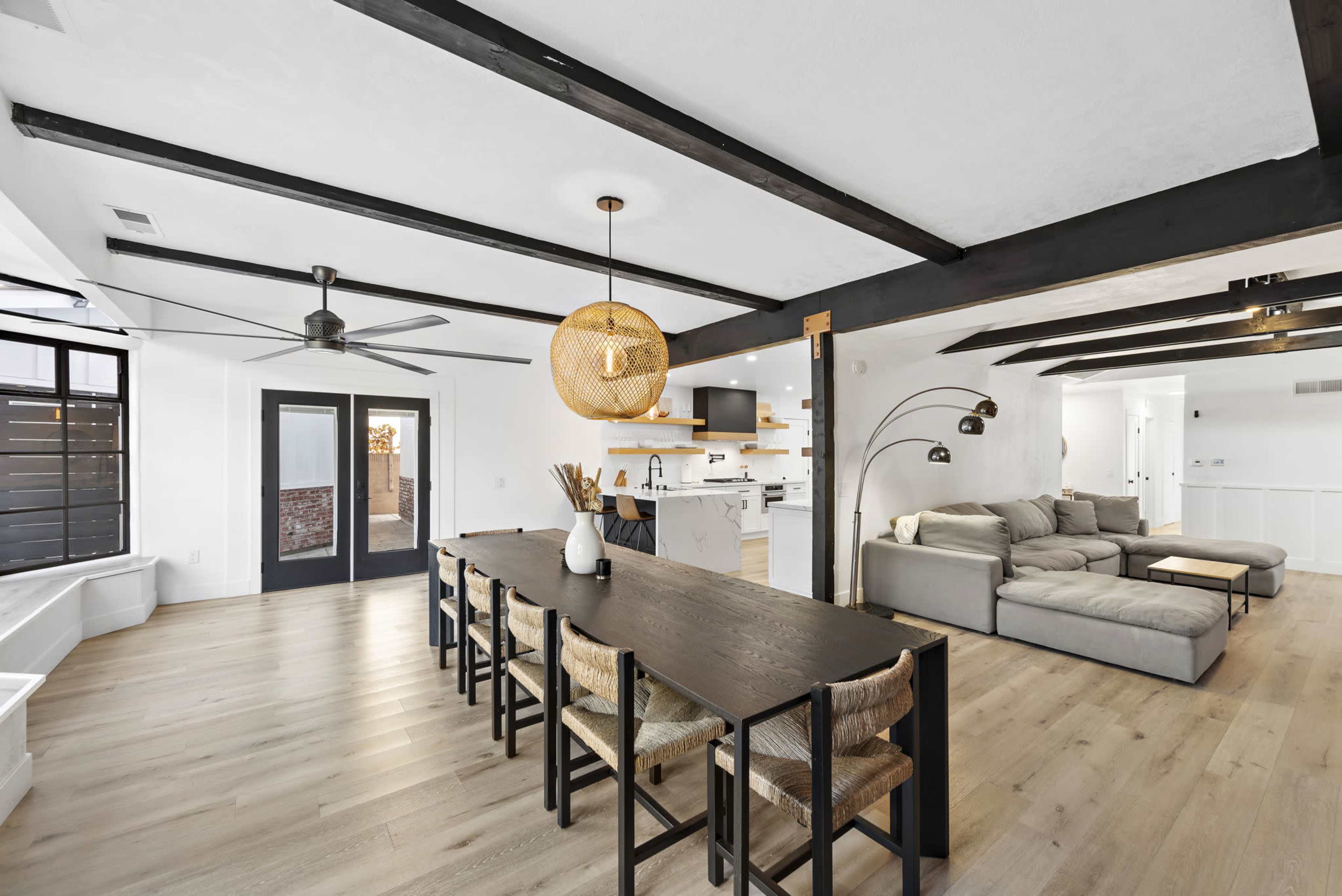 A dining area features a long table surrounded by chairs, leading into a modern kitchen with a large island and a living space with a sectional sofa.