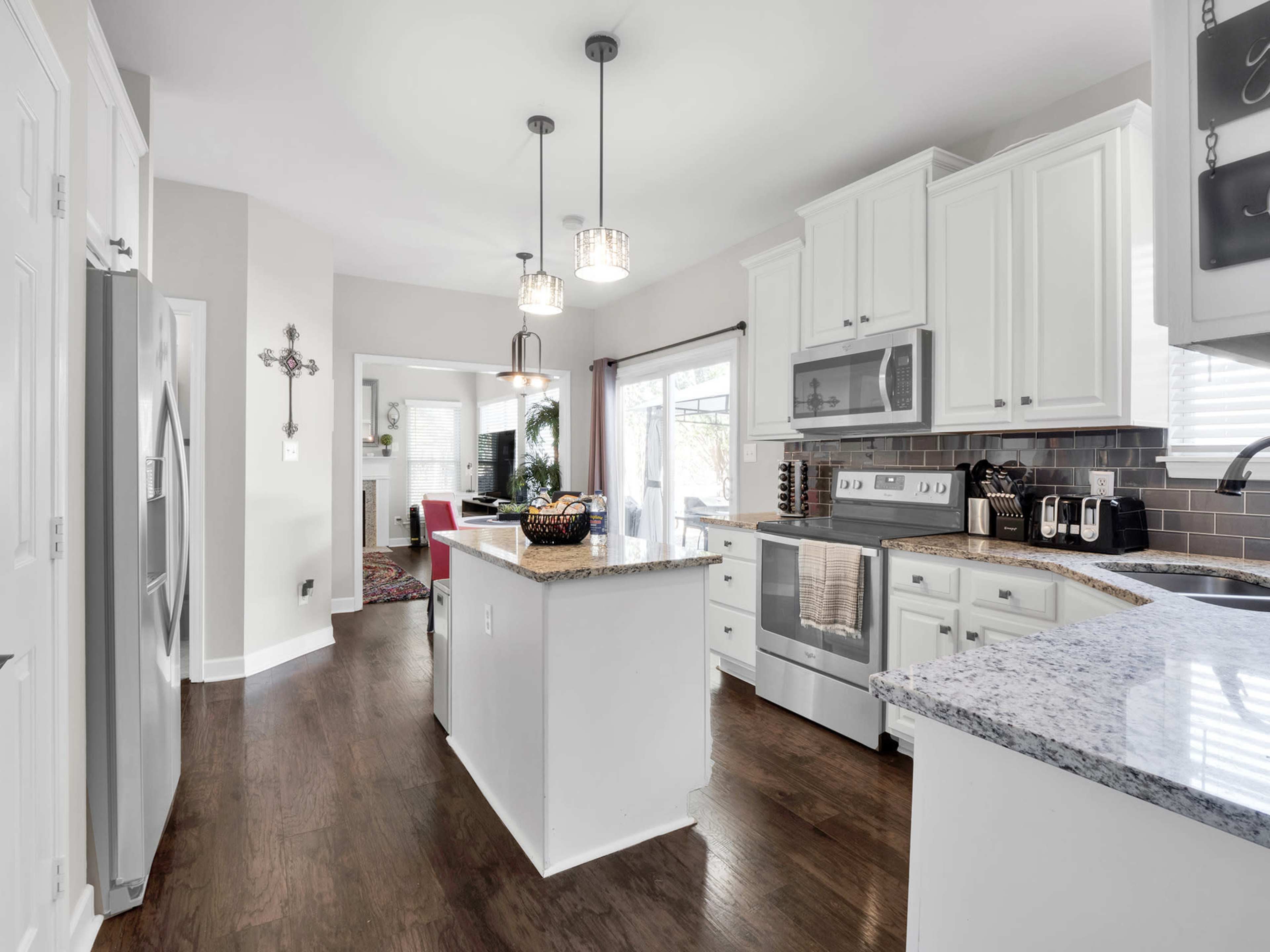 A modern kitchen features white cabinets, a granite island, stainless steel appliances, and a dining area visible in the background.
