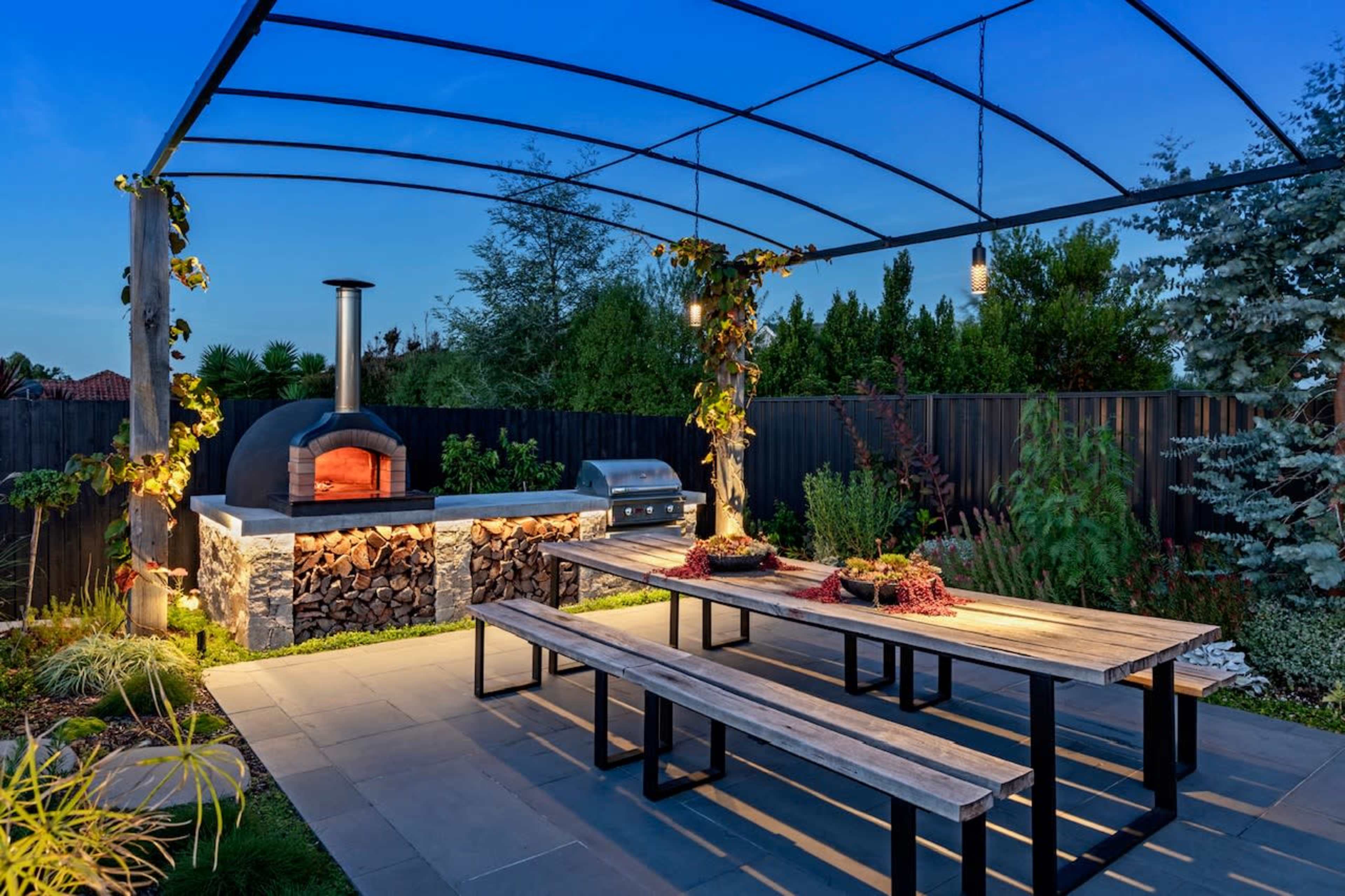 An outdoor patio area with a wood-fired pizza oven, barbecue grill, and a long dining table under a canopy, surrounded by greenery.