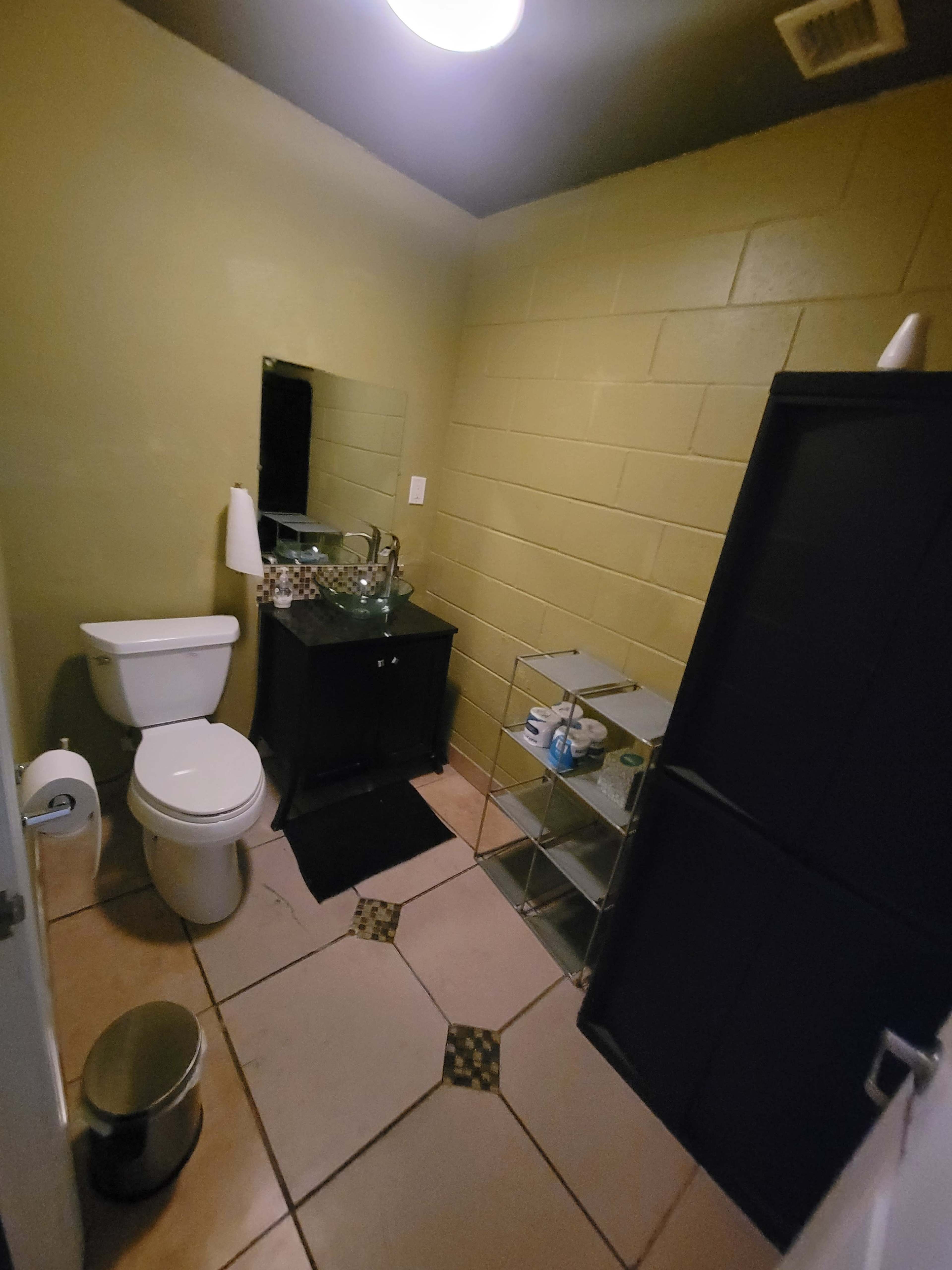 The image shows a small bathroom featuring a toilet, a sink with a cabinet, a mirror, and a shower stall, with beige walls and tiled flooring.