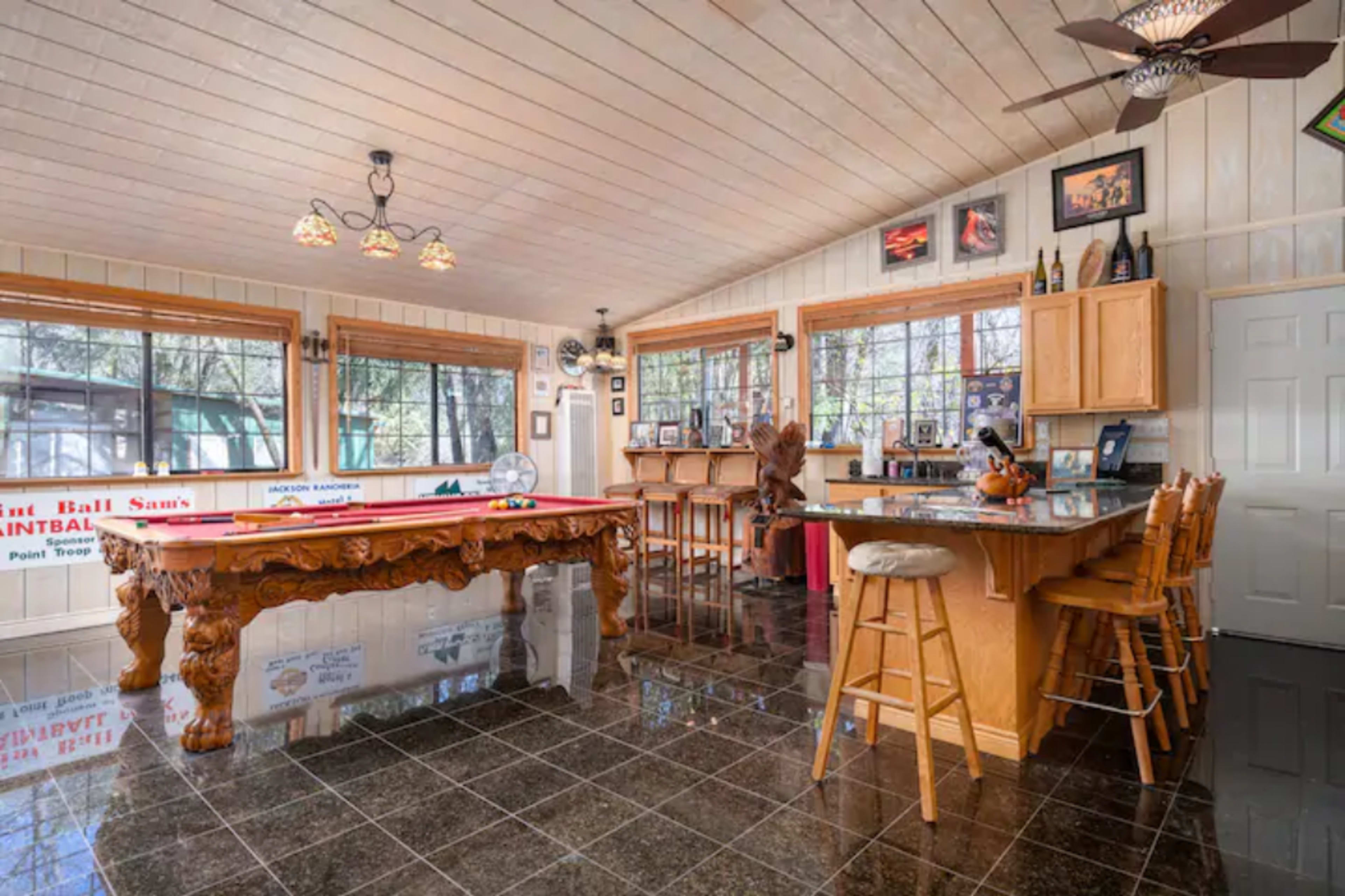 The room features a bar area with wooden stools, a pool table, and large windows that allow natural light to fill the space.