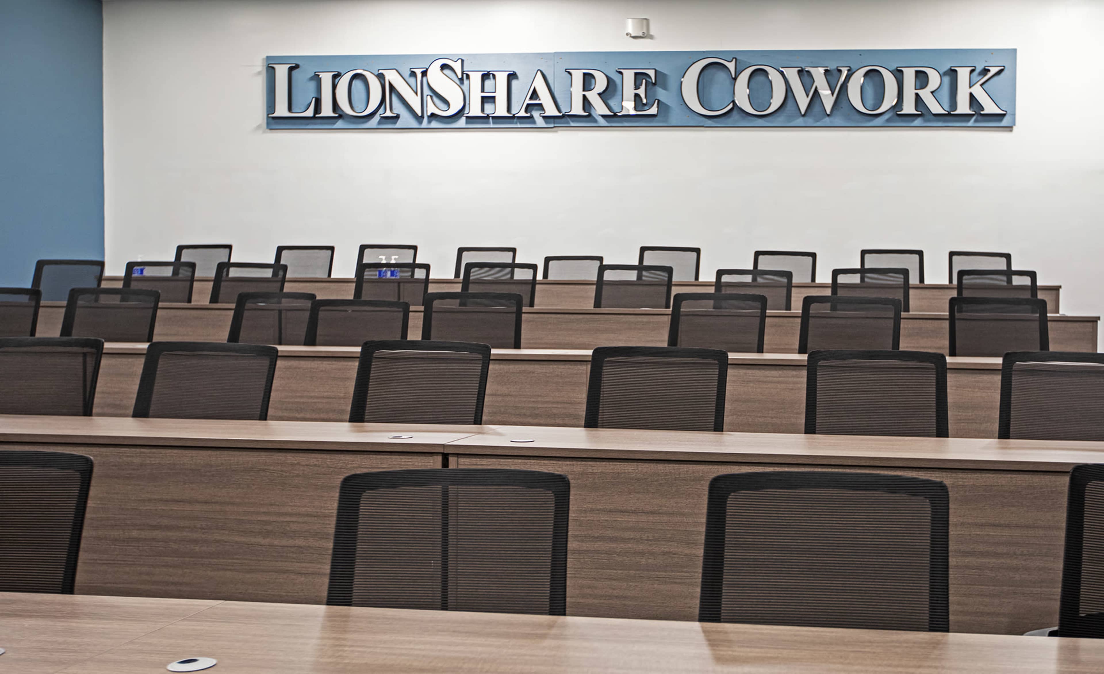 A spacious coworking area with multiple rows of wooden desks and black mesh chairs, featuring a sign that reads "LionShare Cowork."