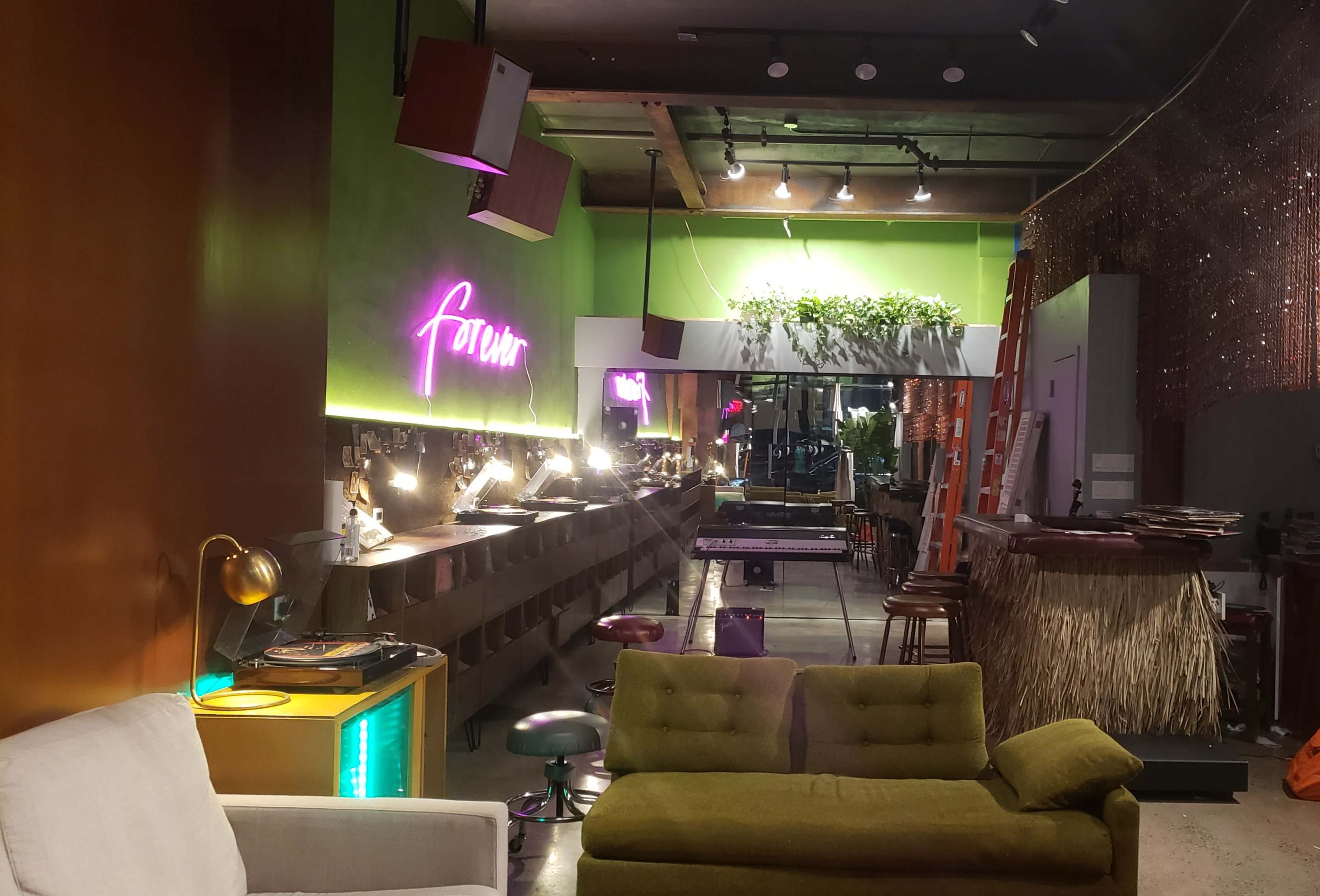 A modern bar interior with green walls, a neon sign, various seating arrangements, and a section adorned with plants.