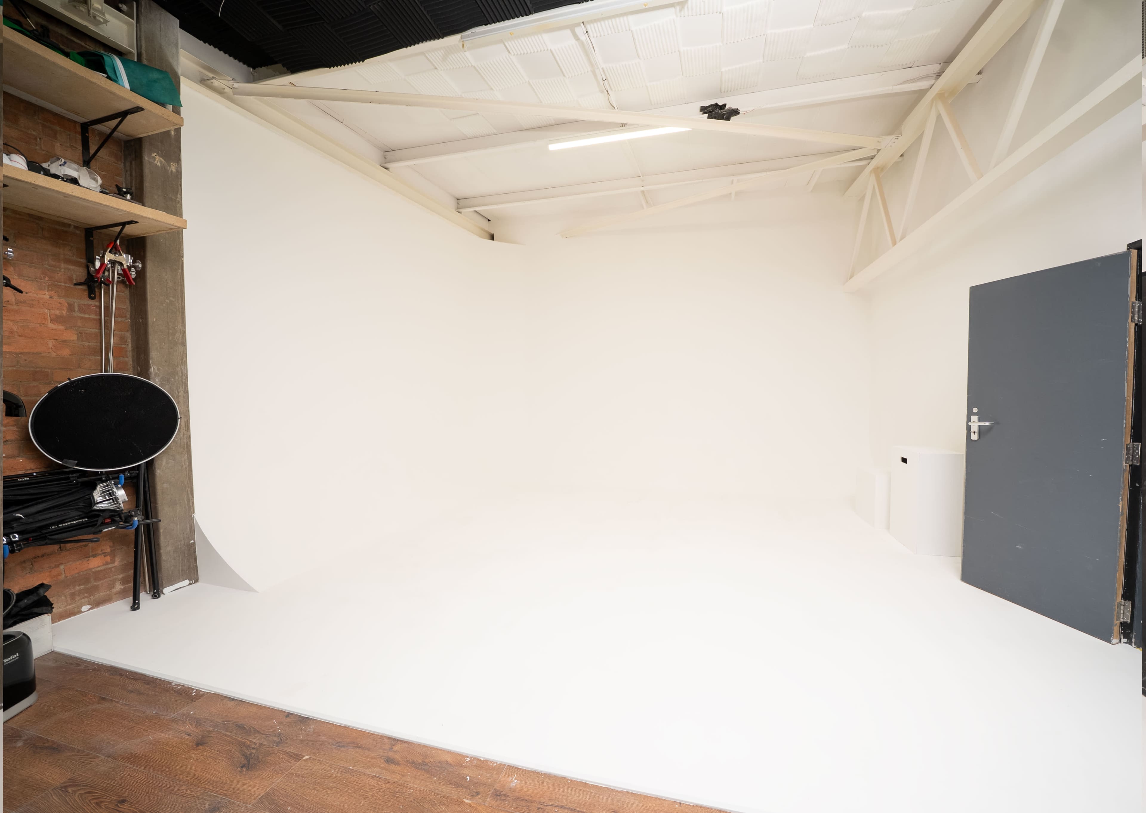 The image shows a spacious, empty studio with white walls, a wooden floor, and a gray door.