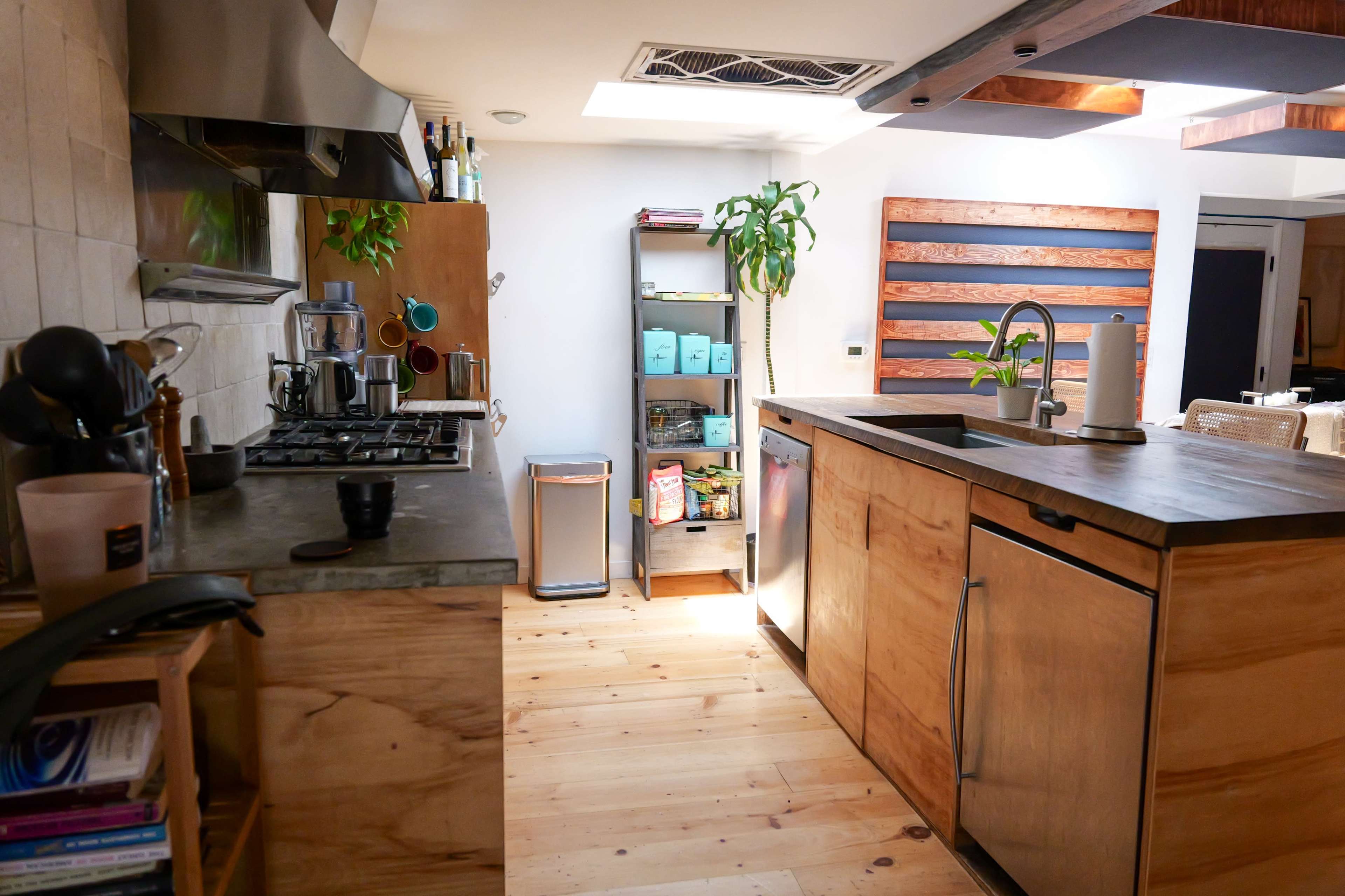 A modern kitchen with wooden cabinets, a concrete countertop, a gas stove, and a shelving unit displaying containers and plants.
