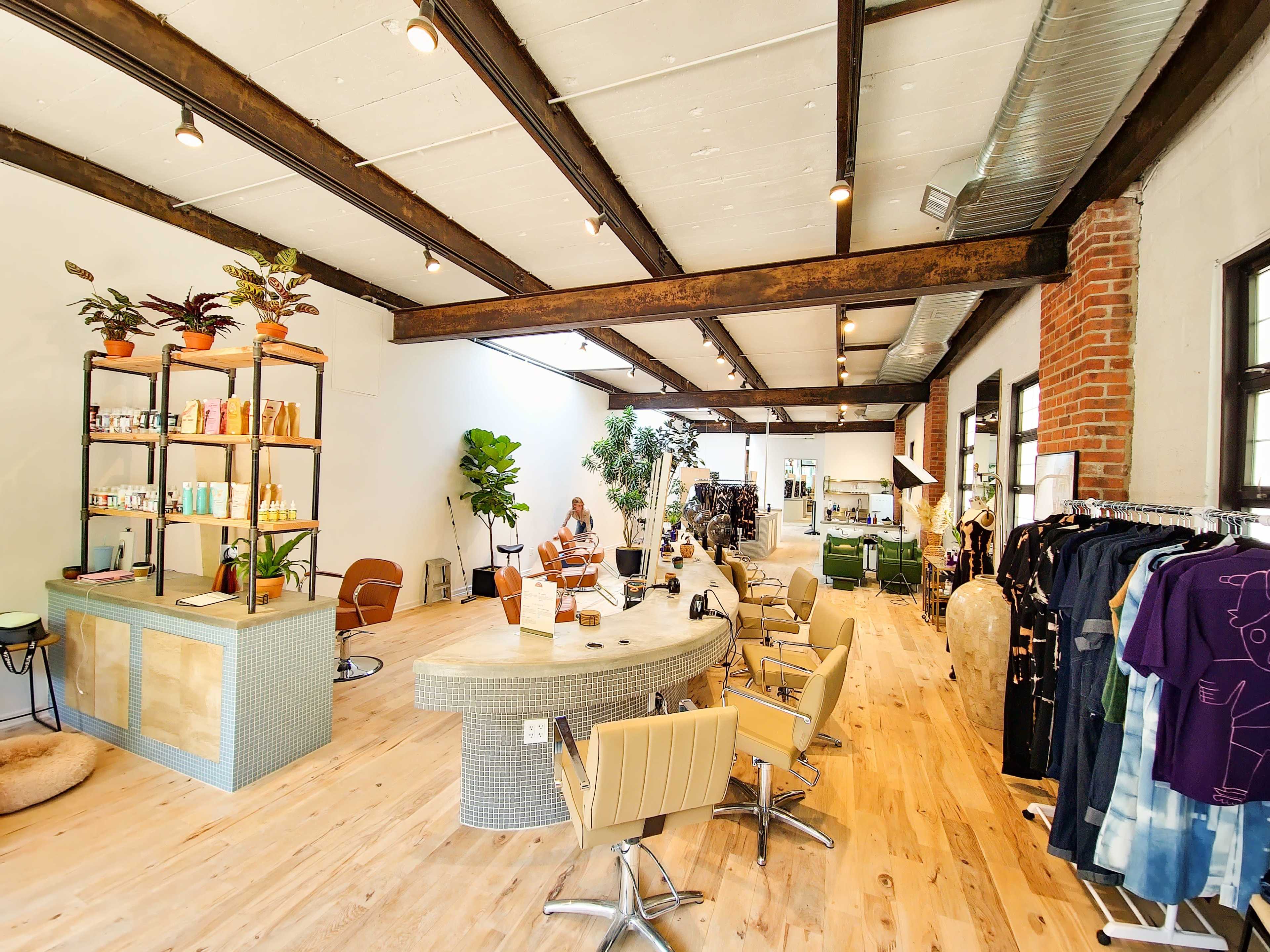 A modern salon interior with a circular reception desk, several styling chairs, plants, and various products displayed on shelves.