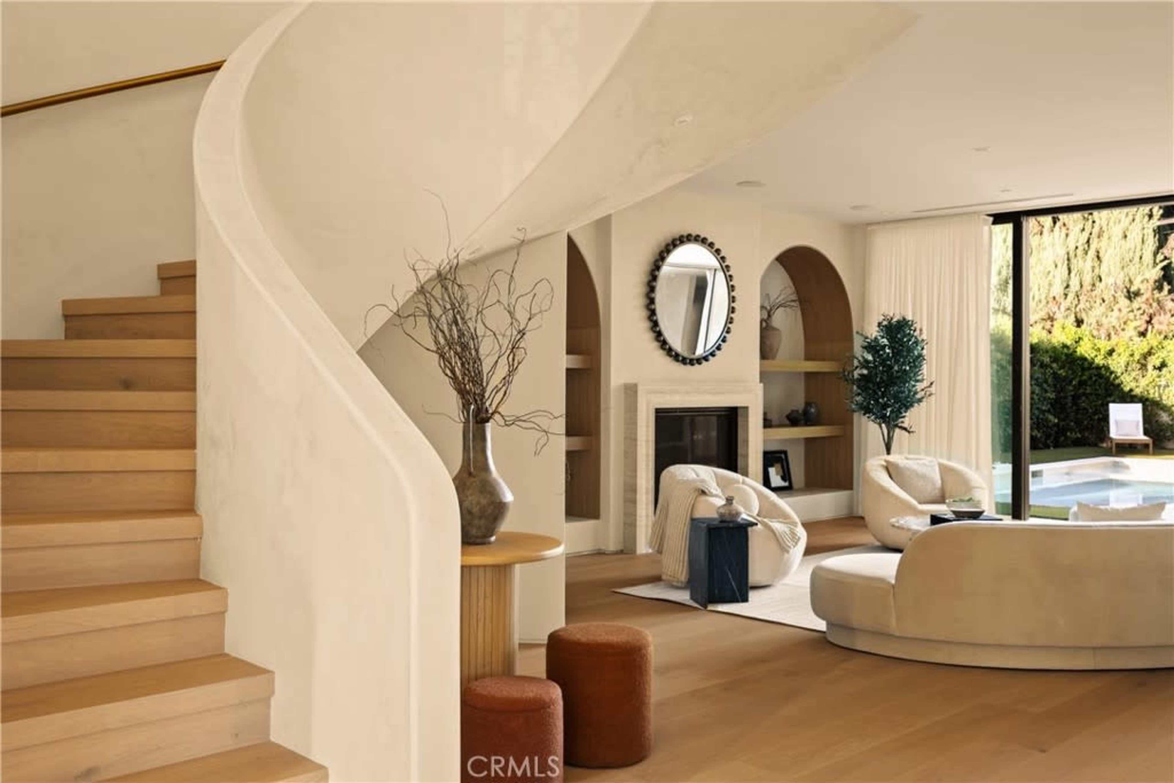 The image shows a modern interior featuring a staircase with a curved railing, a living area with a round mirror, a cozy seating arrangement, and a view of an outdoor space.