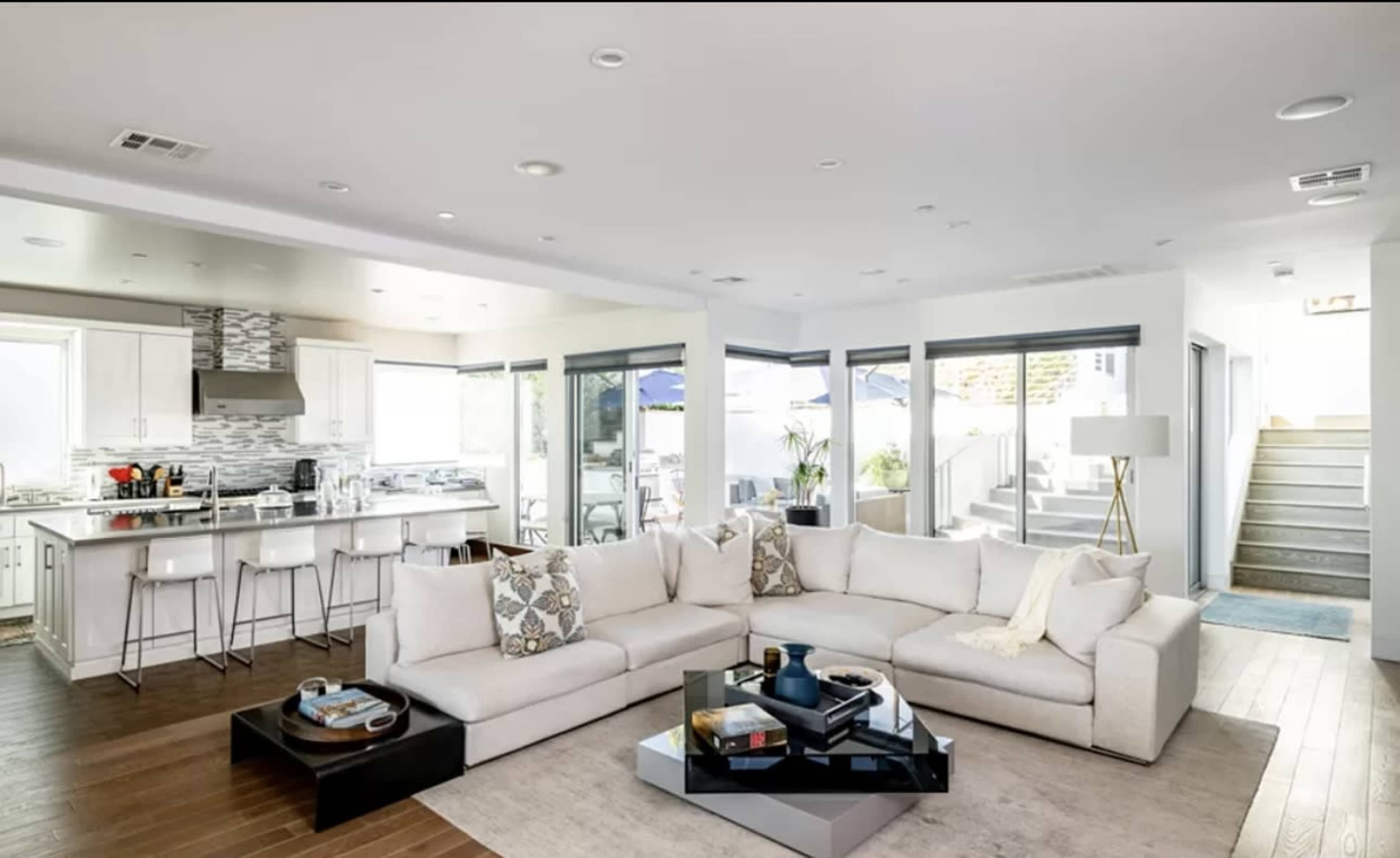 A modern living room featuring a large sectional sofa, a glass coffee table, and an open layout that connects to a kitchen with white cabinetry and stainless steel appliances.