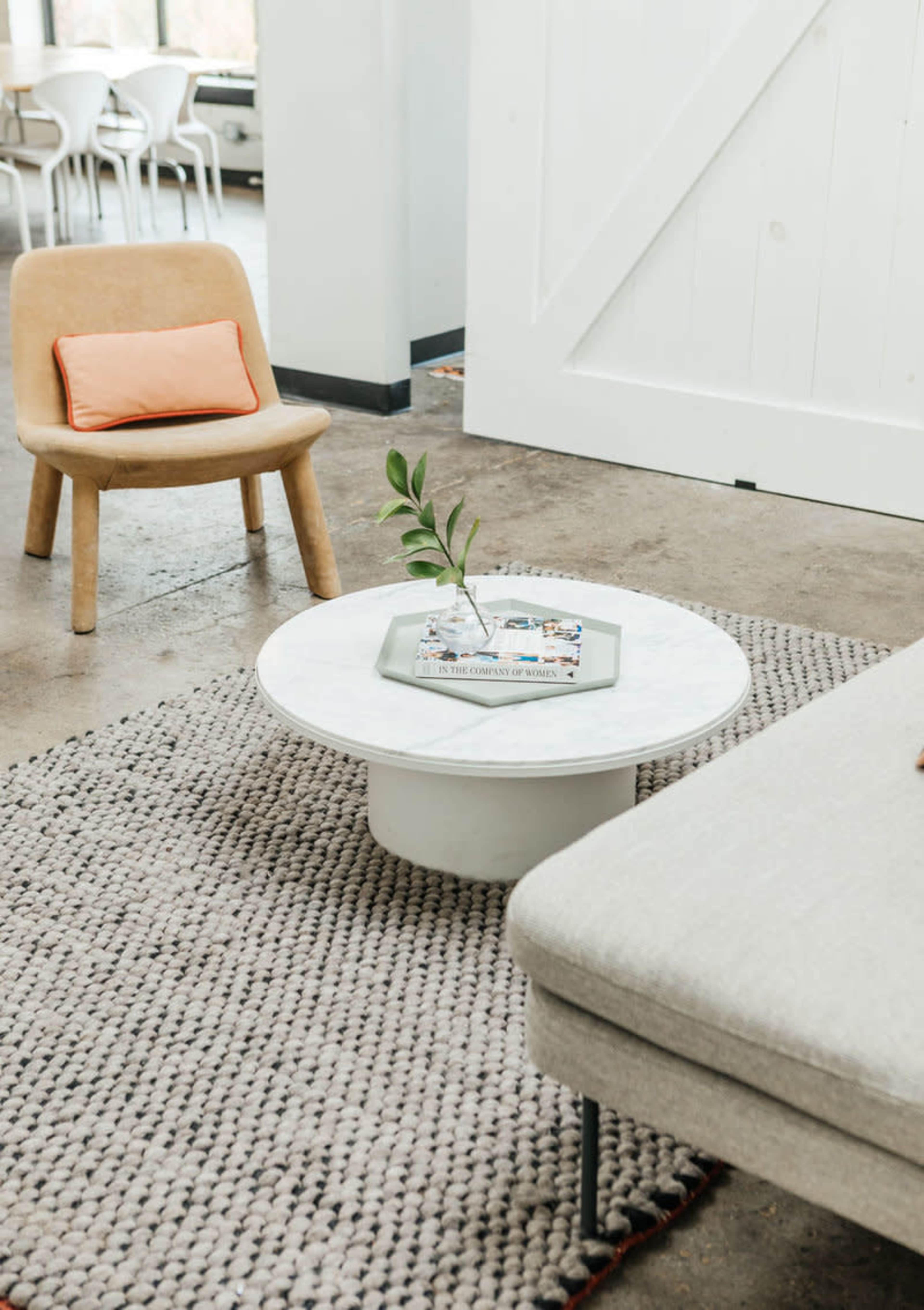 A modern living room features a round marble coffee table atop a textured rug, accompanied by a beige chair and a light gray sofa.