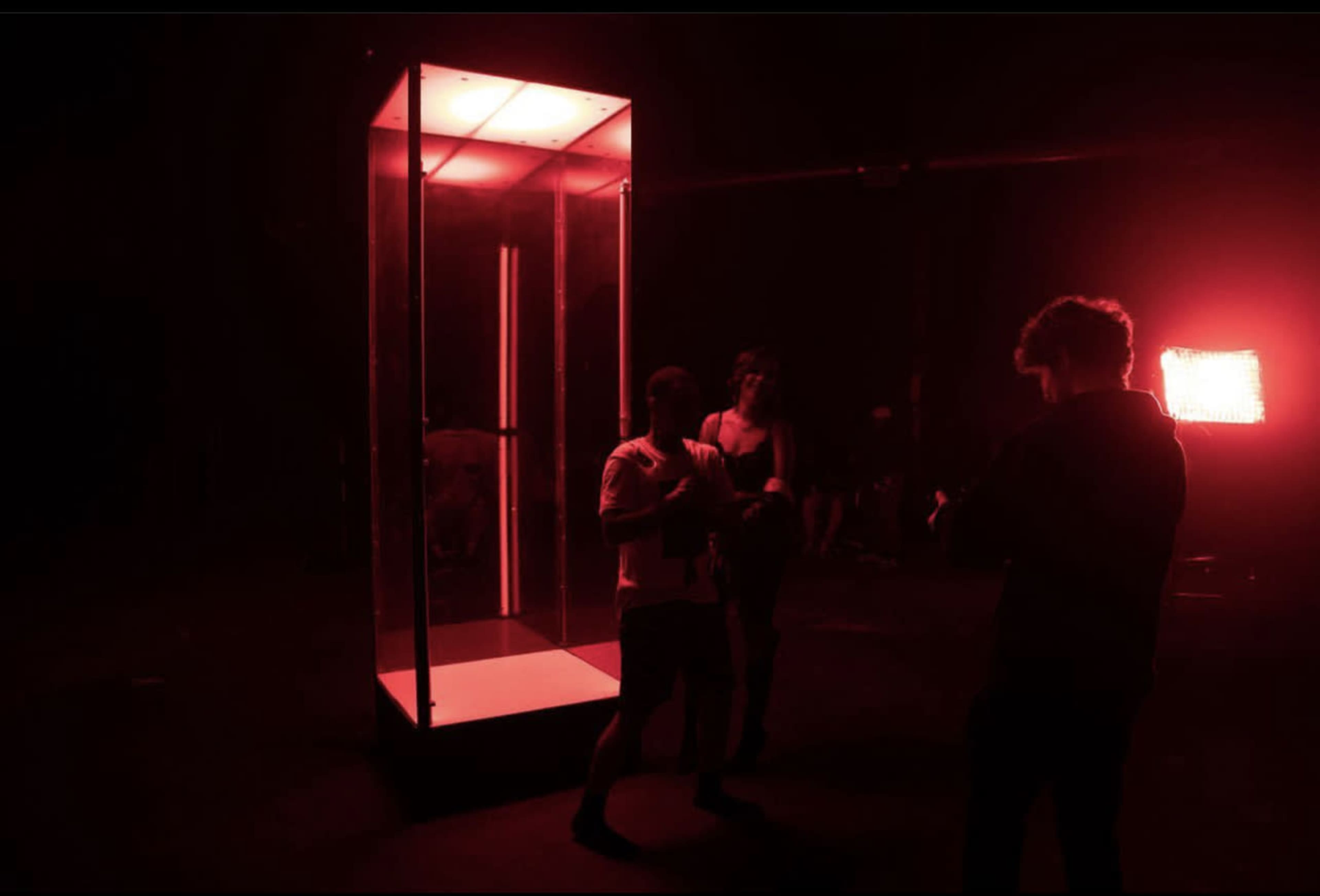 Two people stand near a glass structure illuminated by red lights in a dimly lit space.