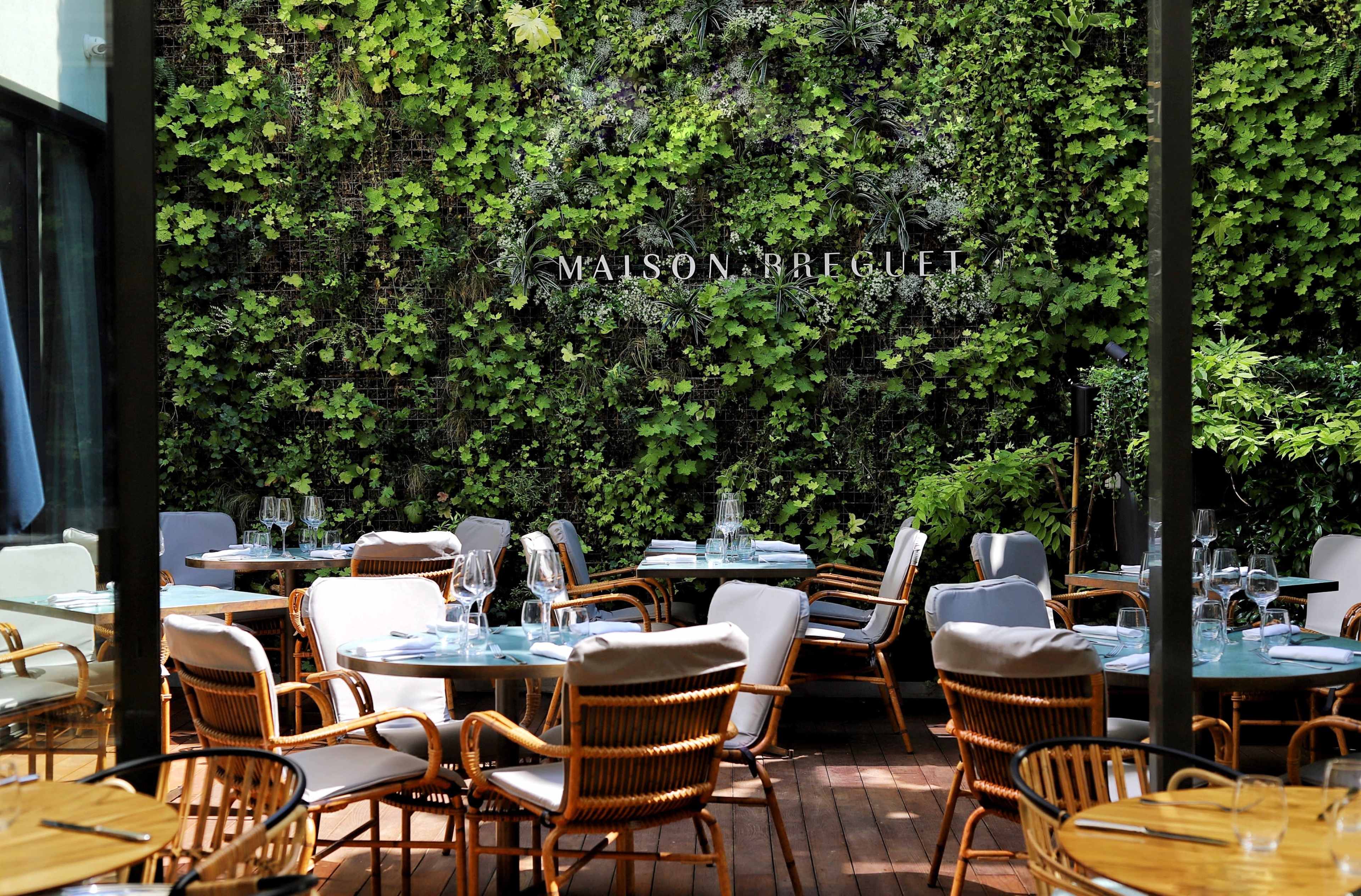 A dining area with wooden tables and chairs is set up against a wall adorned with greenery, featuring the name "MAISON BREGUET" prominently displayed.