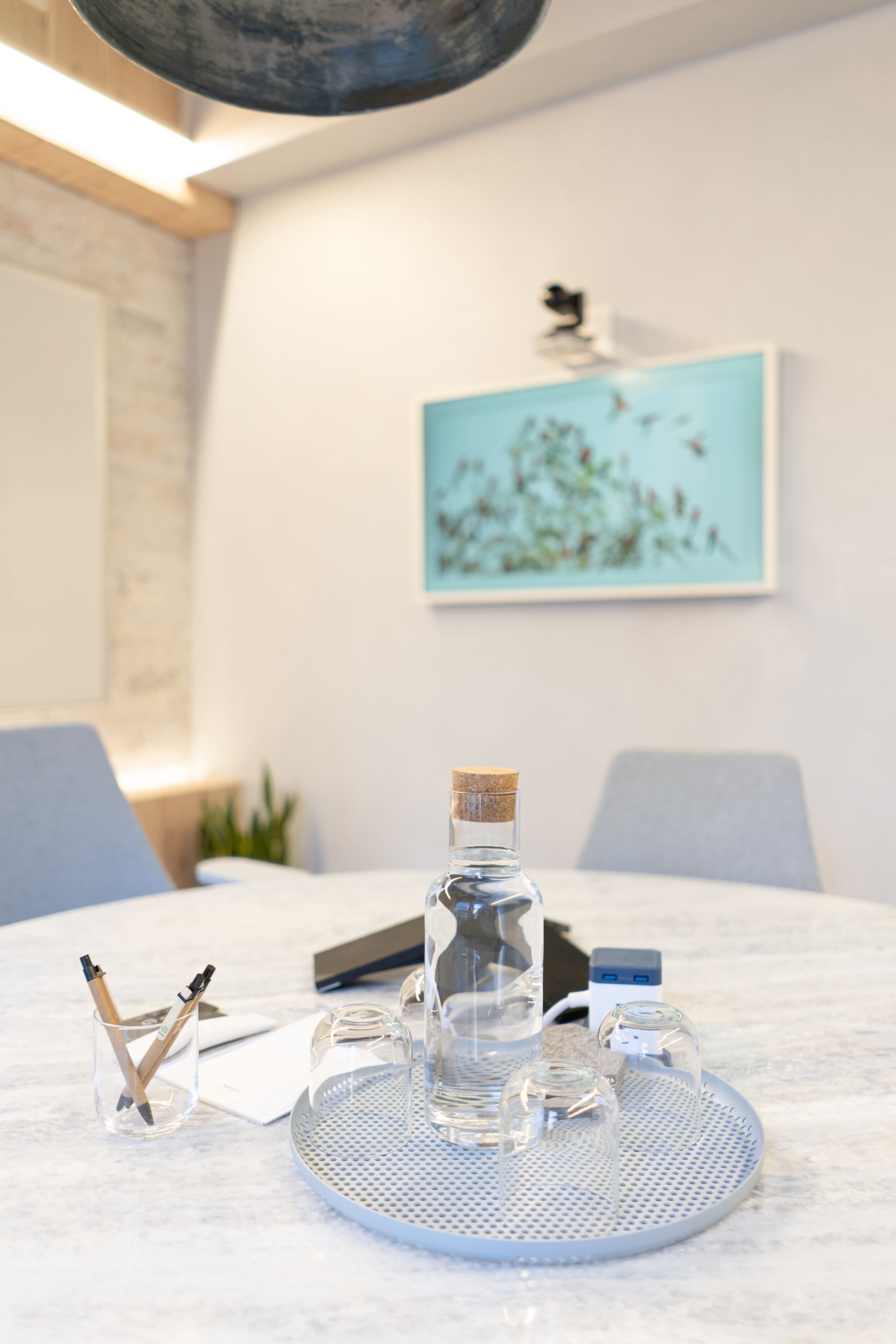 A clear glass water bottle and two small glasses are placed on a round tray in a modern meeting room with light-colored furniture and a piece of artwork on the wall.