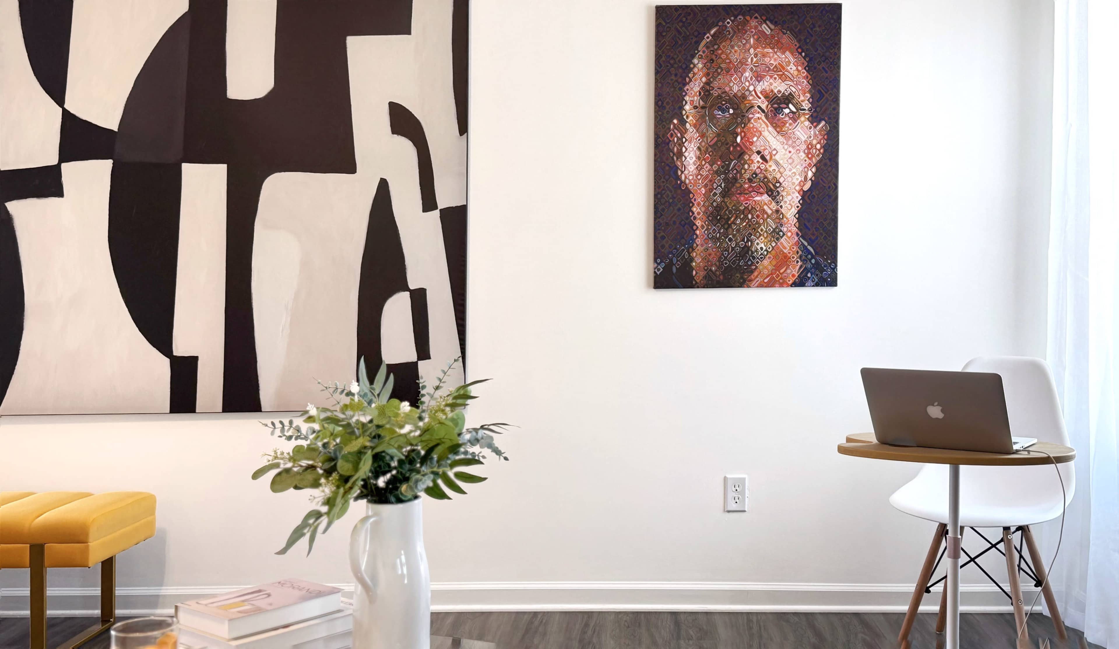 The image shows a modern interior featuring a large abstract painting on the wall, a framed portrait hanging above a small table with a laptop, and a vase of flowers in the foreground.