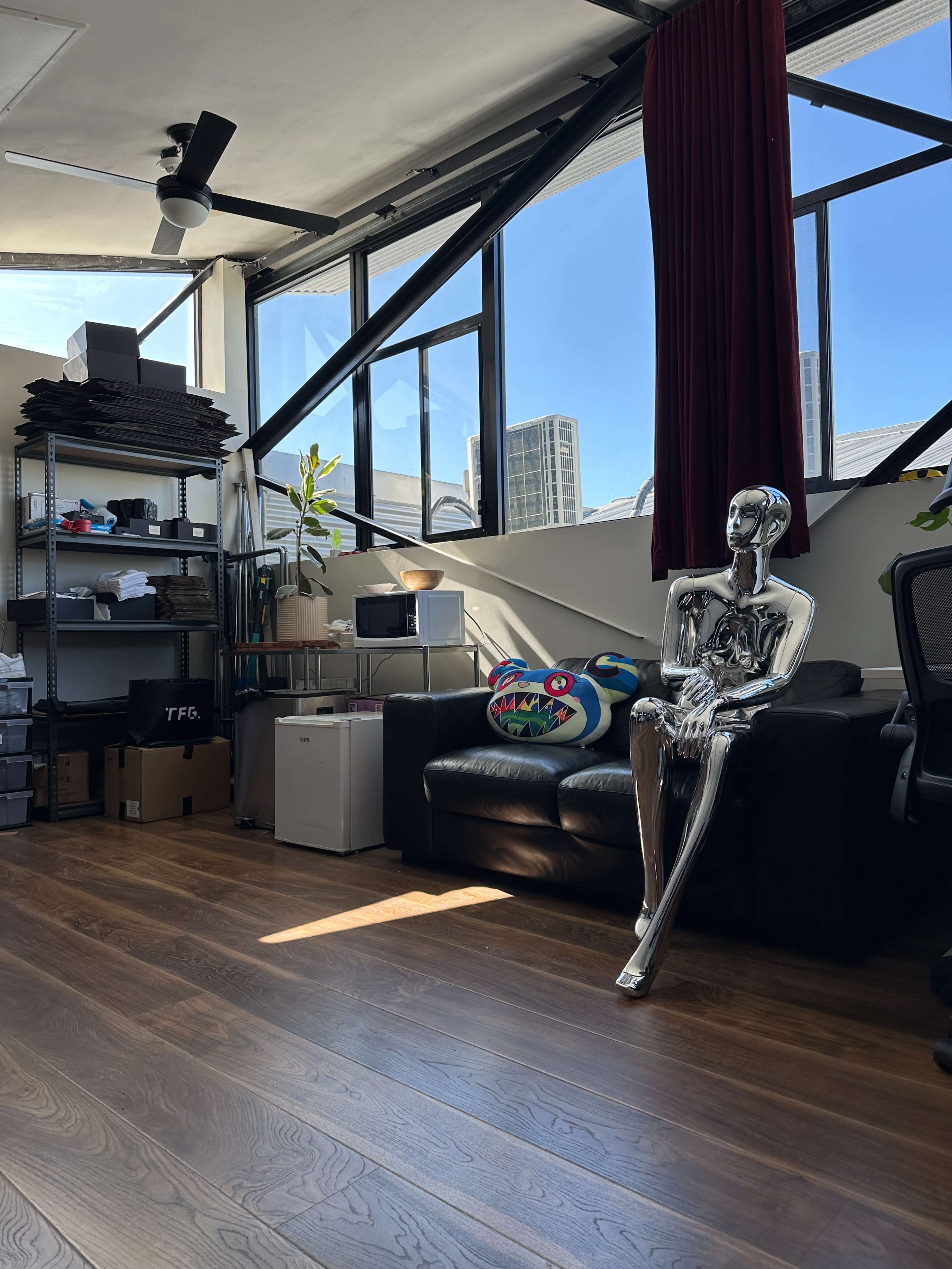 The image shows a modern office space with a black couch, a chrome mannequin seated beside it, and shelves filled with various items in the background.