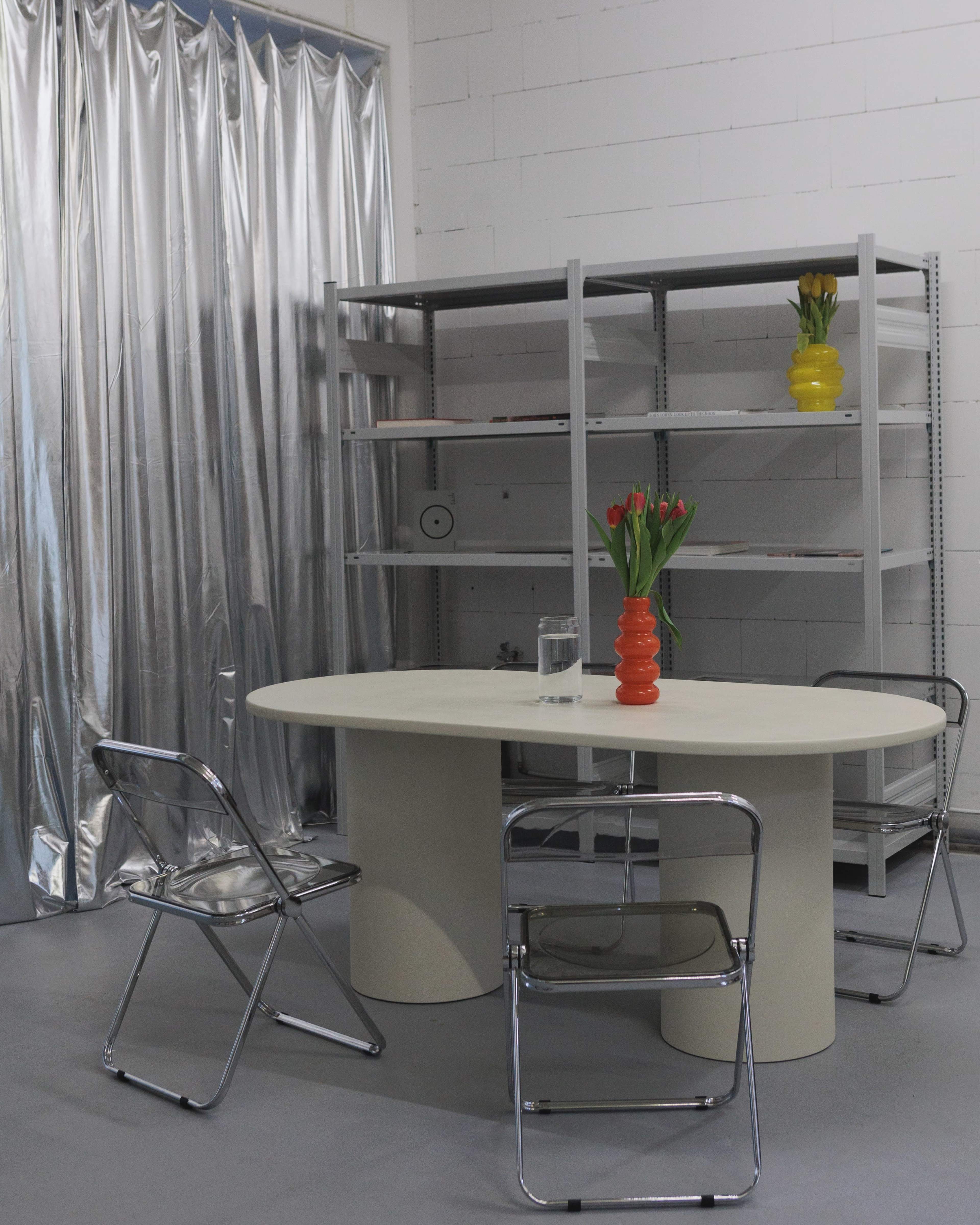 A round table with a beige surface is situated in a room with silver sheeting and a metal shelving unit, accompanied by three transparent chairs and a vase of tulips.
