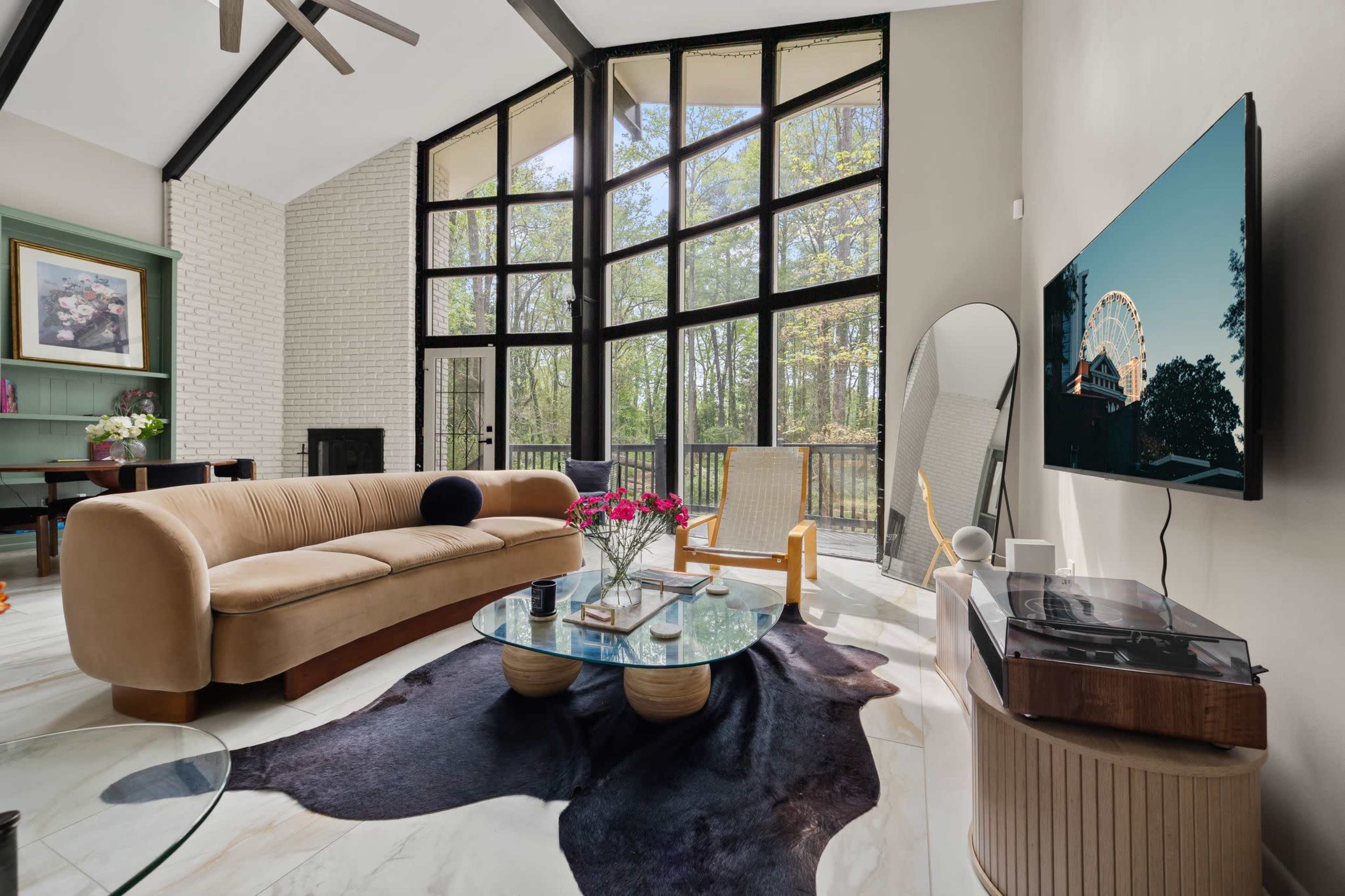 A modern living room features a large glass wall, a curved beige sofa, a round coffee table, and a television mounted on the wall, with sunlight streaming in through the windows.