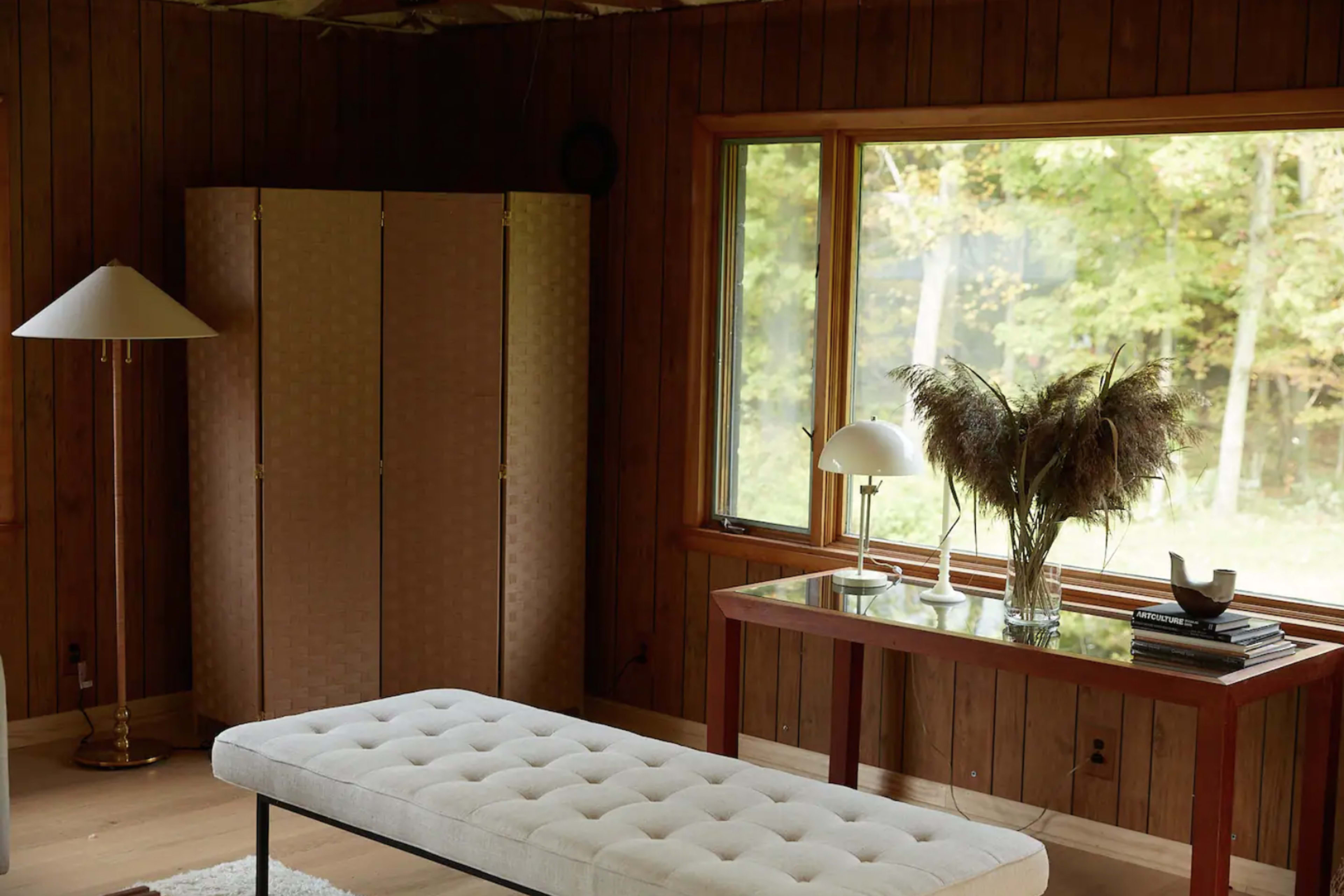 The room features a wooden interior with a bench, a glass-top table with decorative items, and large windows overlooking a forested area.