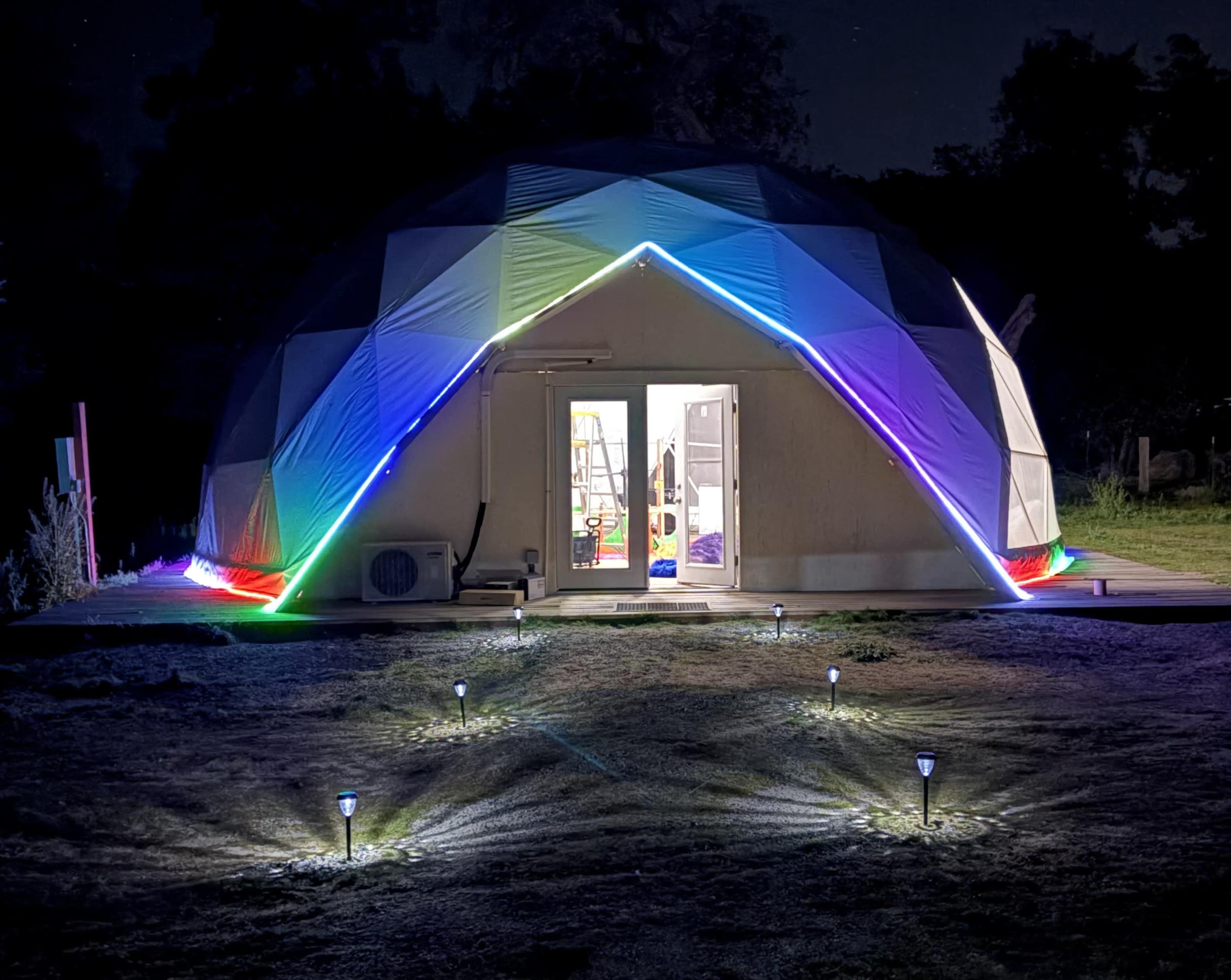 A geodesic dome structure is illuminated with colorful LED lights at night, with landscaped lighting in the foreground.