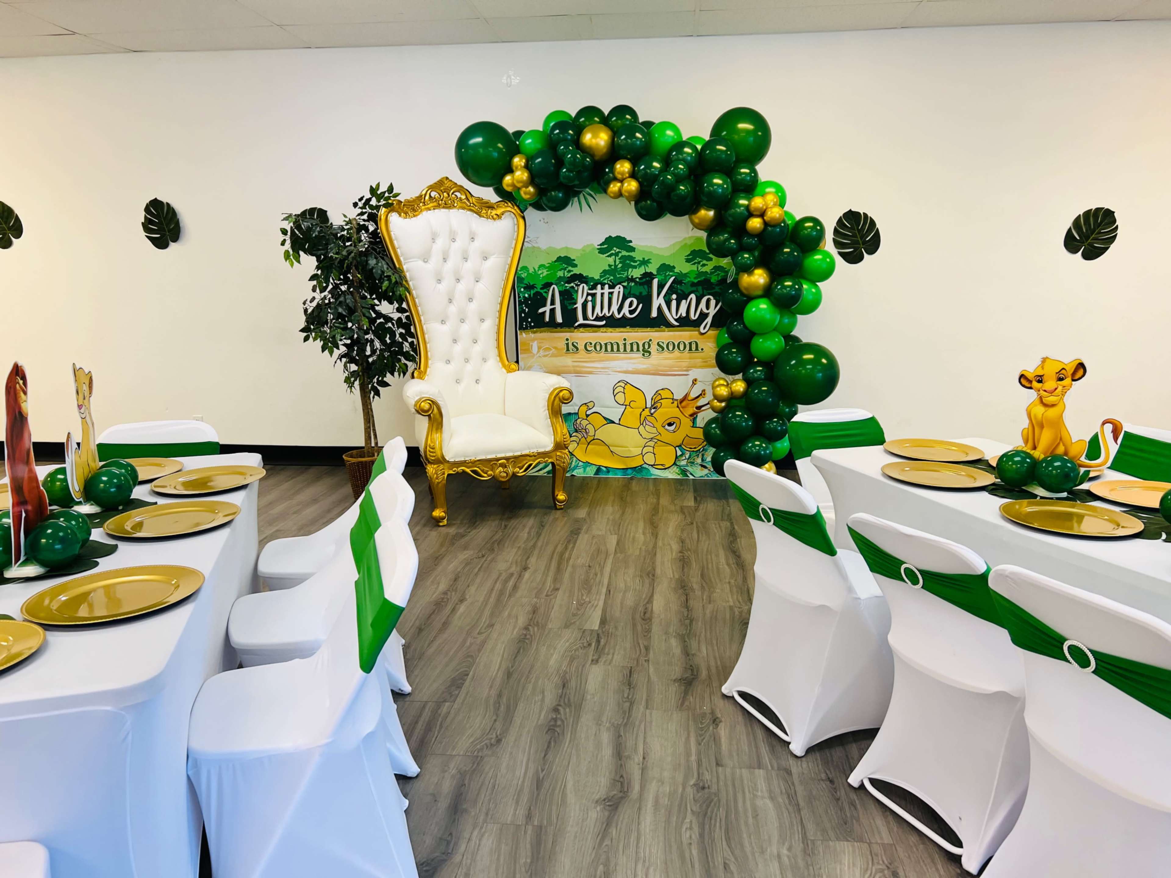 A decorated party venue with white tables set with green accents and gold plates, featuring a throne-like chair surrounded by a balloon arch and a Lion King-themed backdrop.