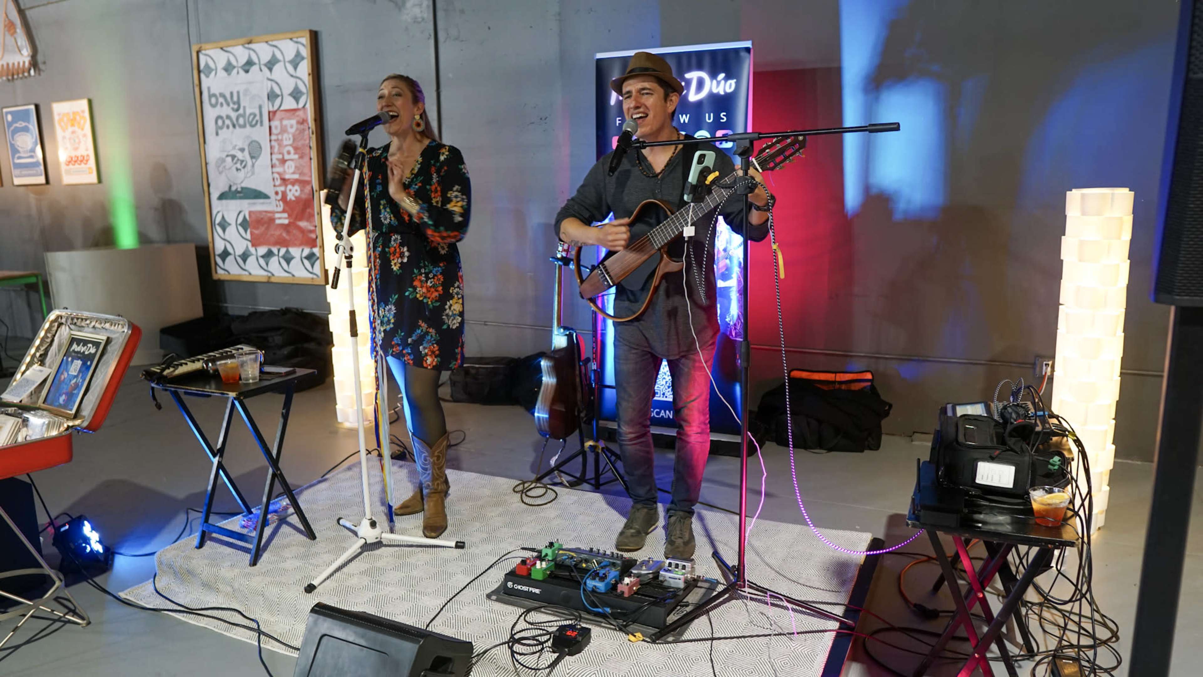 Two musicians perform on stage, one singing into a microphone and the other playing an acoustic guitar, with colorful lights illuminating the backdrop.