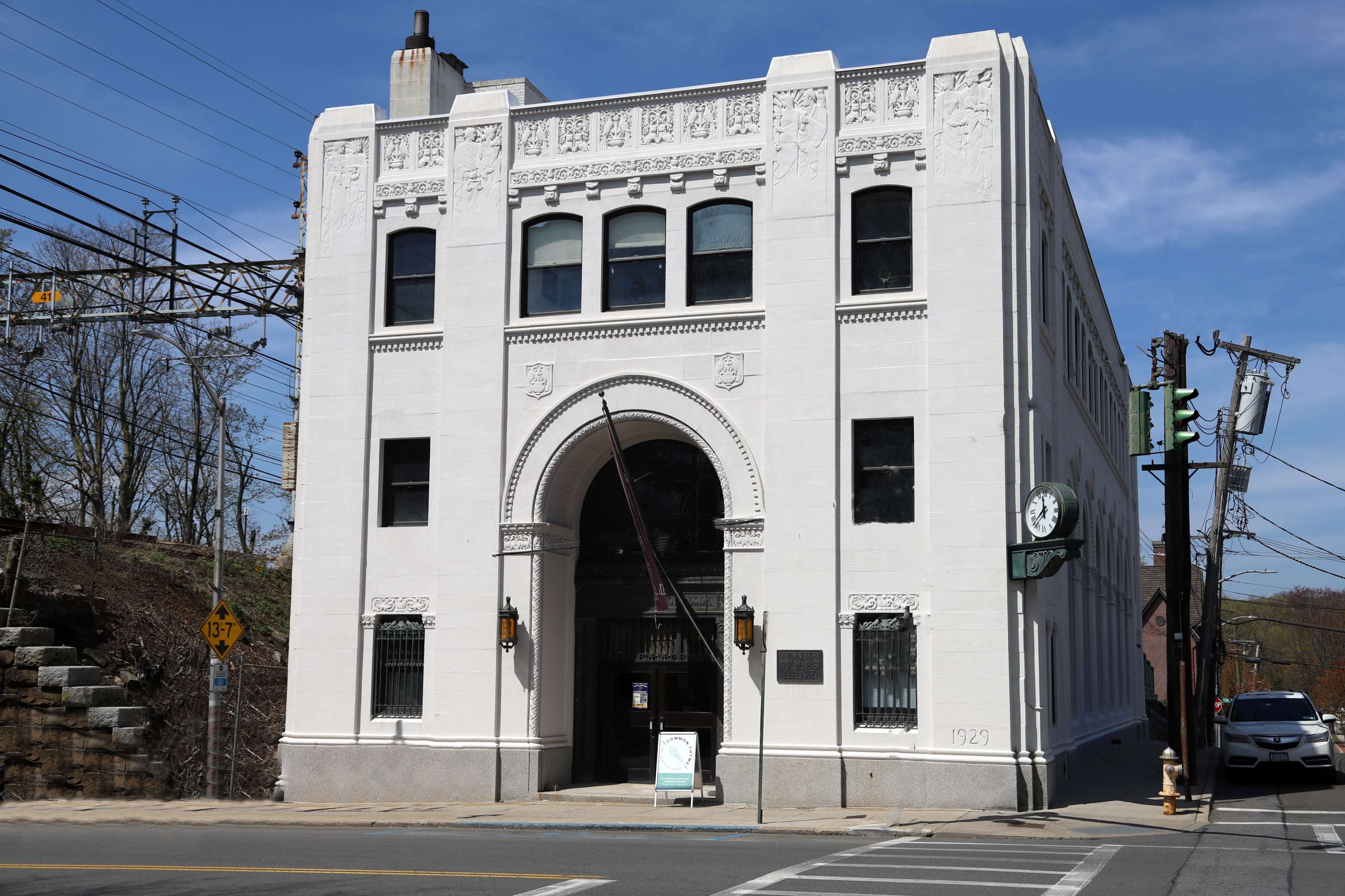 The image shows a white, ornate building with arched windows and a decorative facade, located on a street with utility poles in the background.