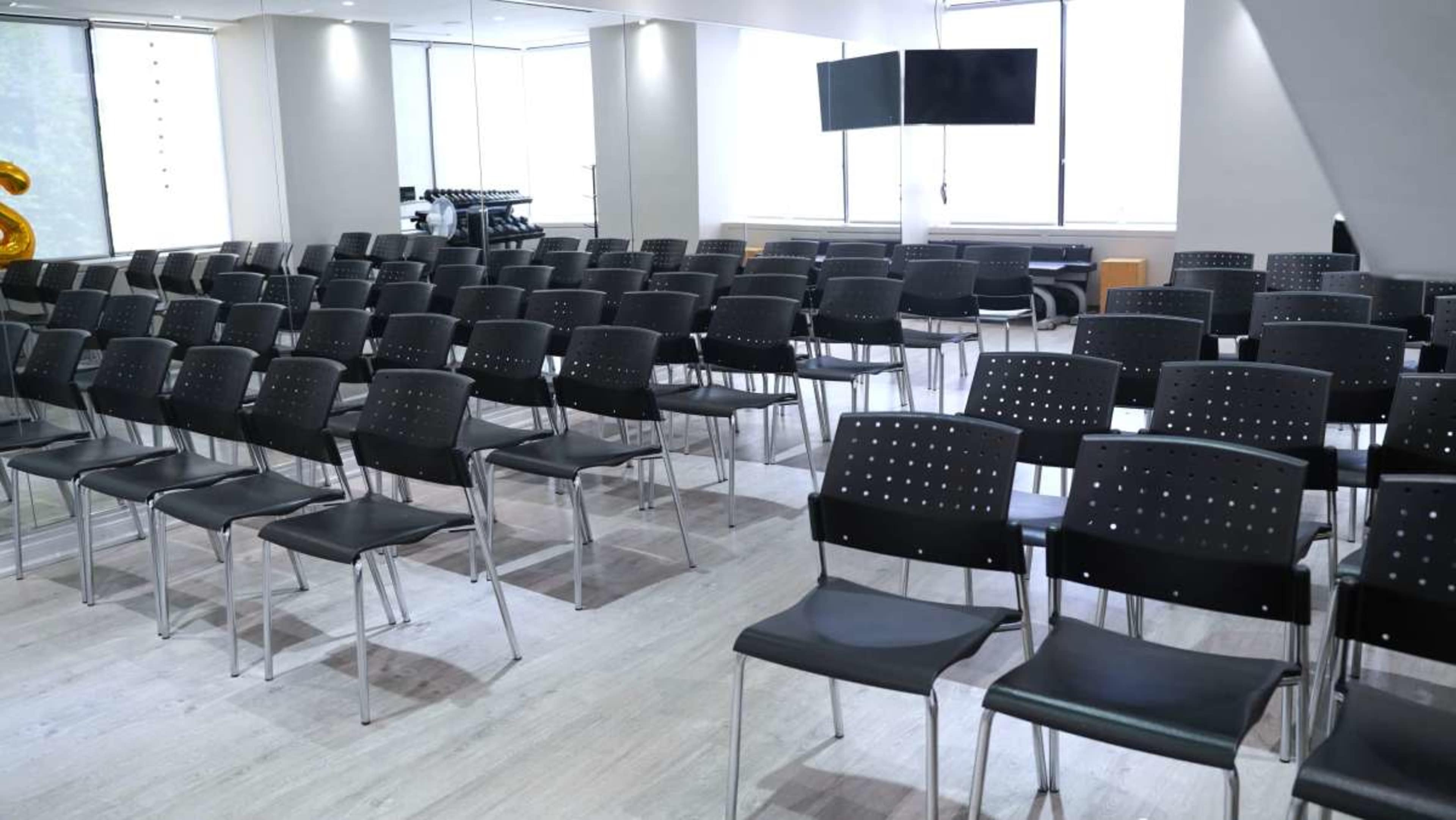 The image shows a row of black chairs set up in a modern, well-lit meeting room, with a small stage and two large screens visible in the background.