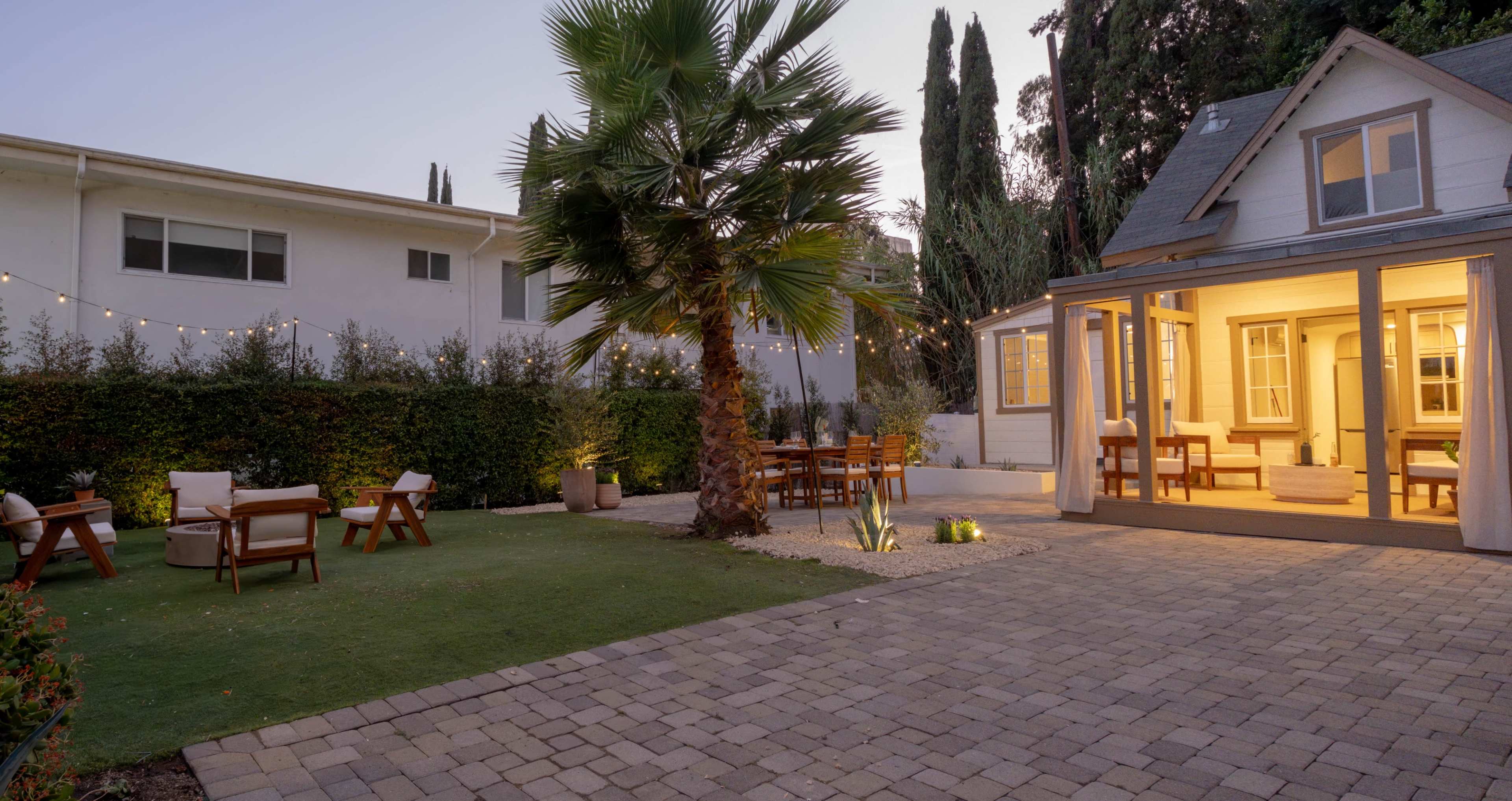 The image shows a backyard with a paved patio, outdoor seating, a palm tree, and a house with illuminated windows surrounded by greenery.
