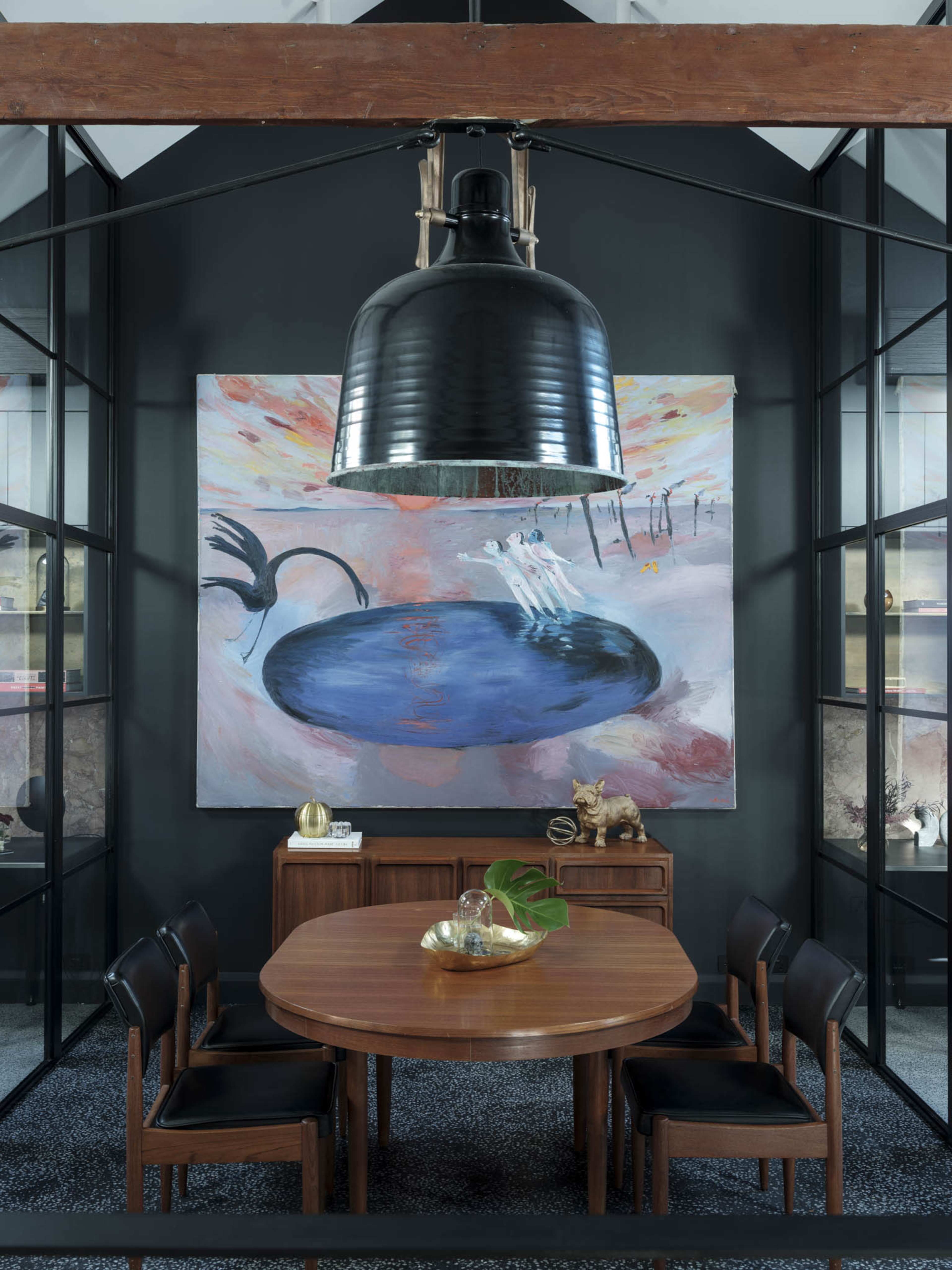 A round wooden table surrounded by black chairs is illuminated by a large pendant light, with a colorful abstract painting on the wall behind it.
