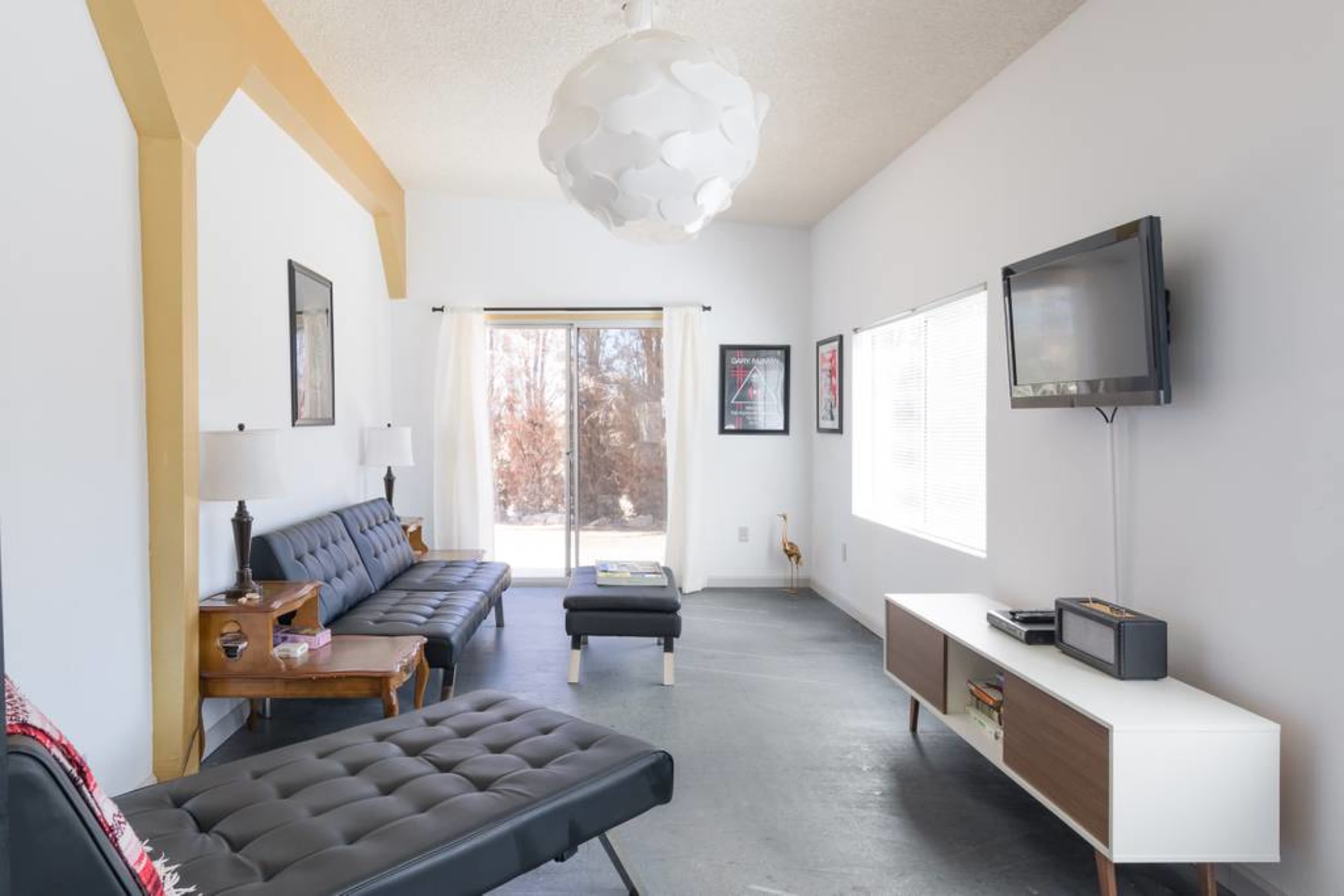 A modern living room features a sleek black sofa, a coffee table, a wall-mounted television, and large windows that provide natural light.