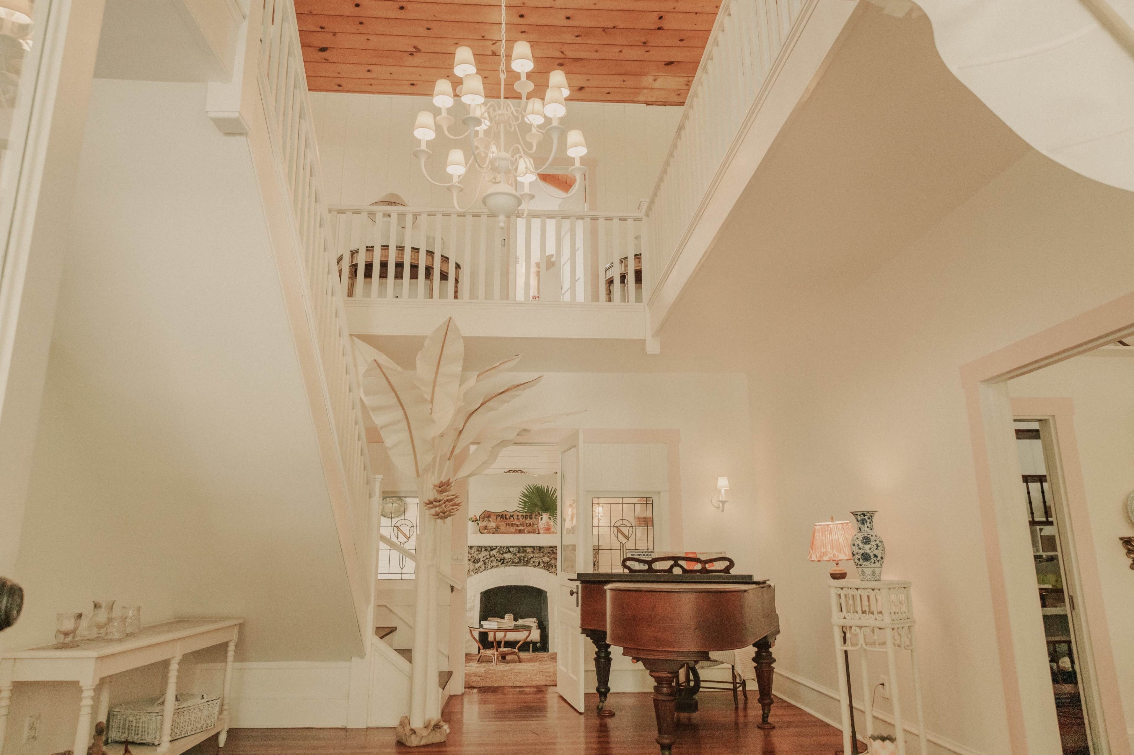 The image shows a light-filled entryway with a staircase, a chandelier, a grand piano, and decorative elements like a large plant and a lamp.