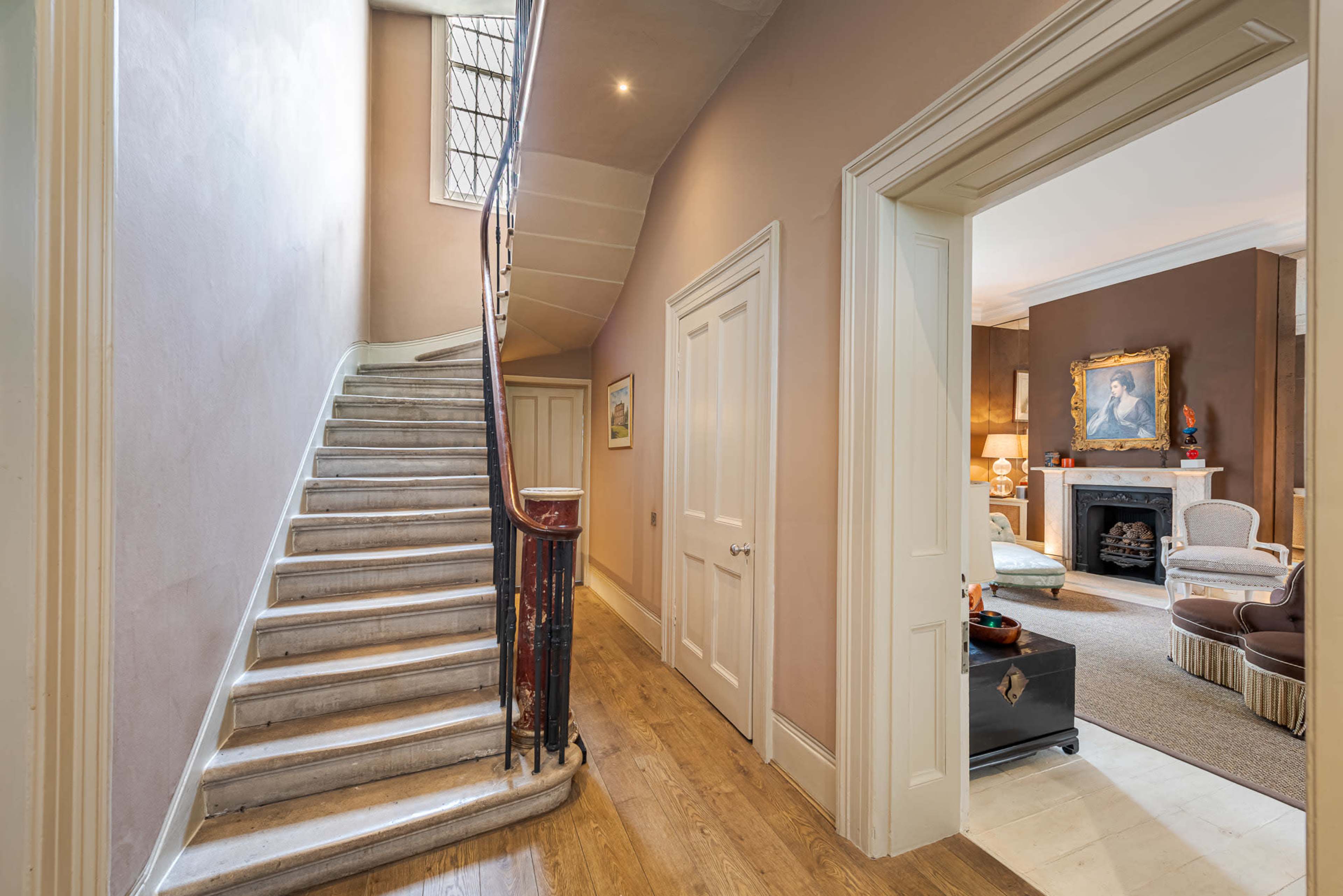 The image shows a hallway leading to a staircase with wooden steps, framed by walls painted in light tones and an open doorway leading to a furnished living room.