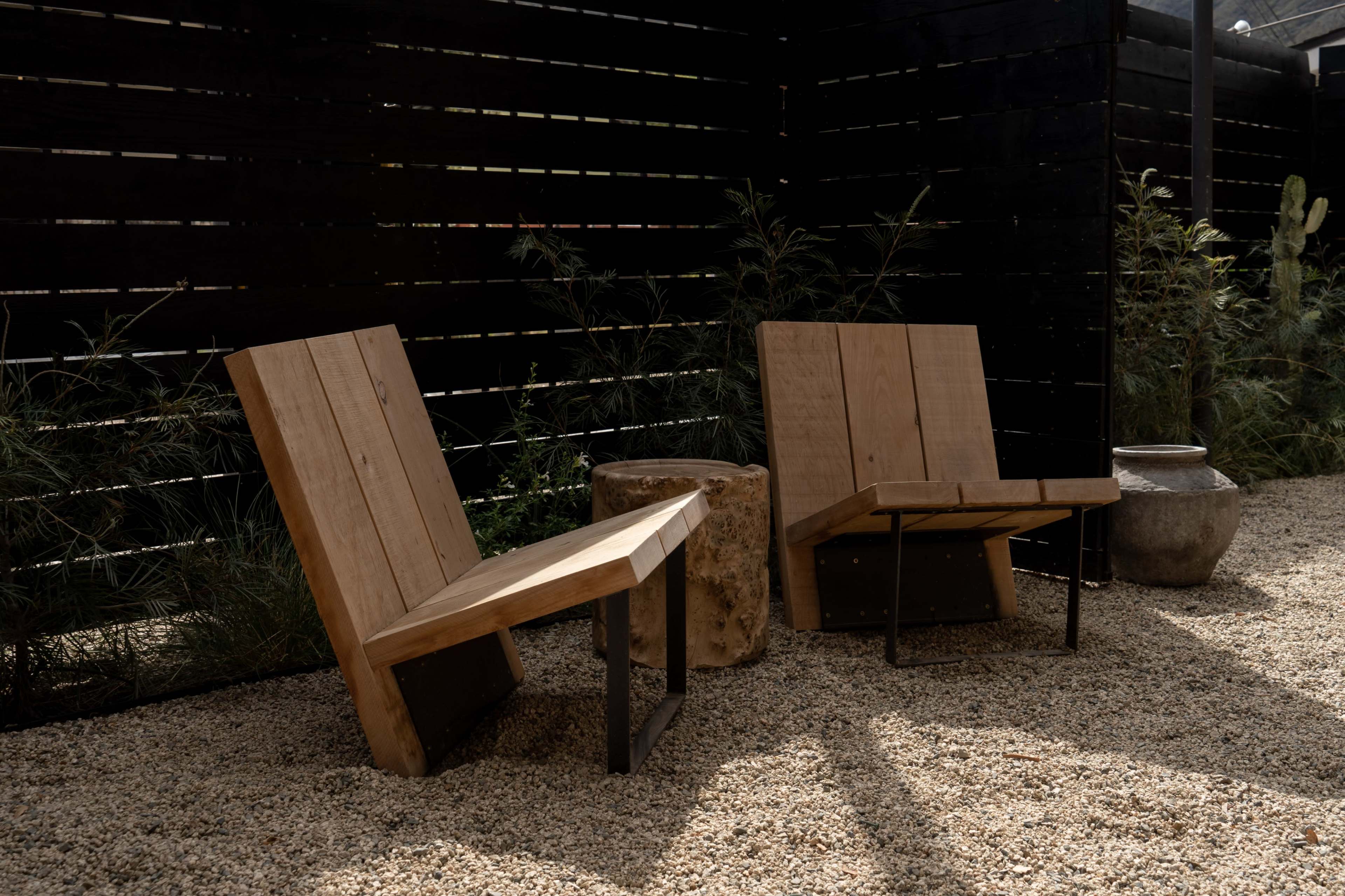 Two wooden chairs with angled backs are positioned on a gravel surface beside a large stone planter and a black wooden fence.