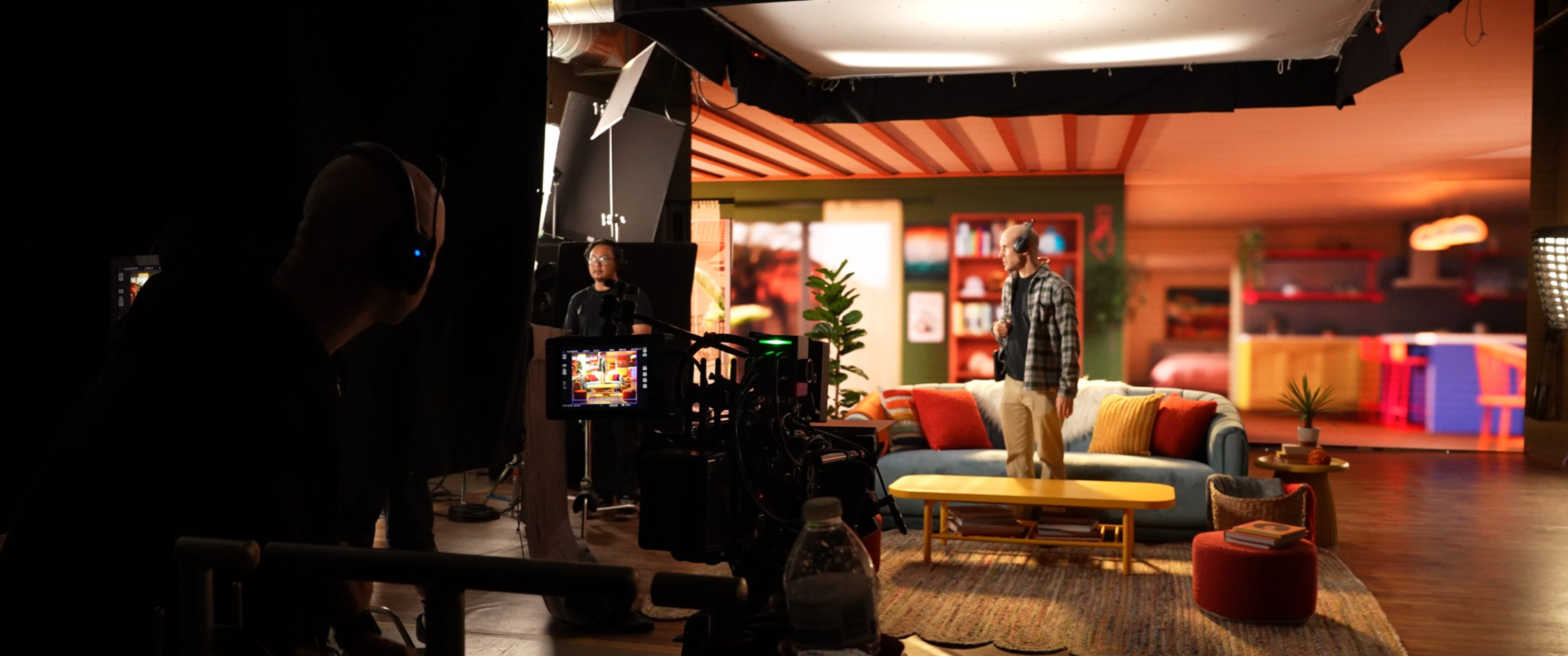 The image shows a film set with a camera crew operating in the foreground and an actor standing on a colorful sofa in a vibrant living room backdrop.