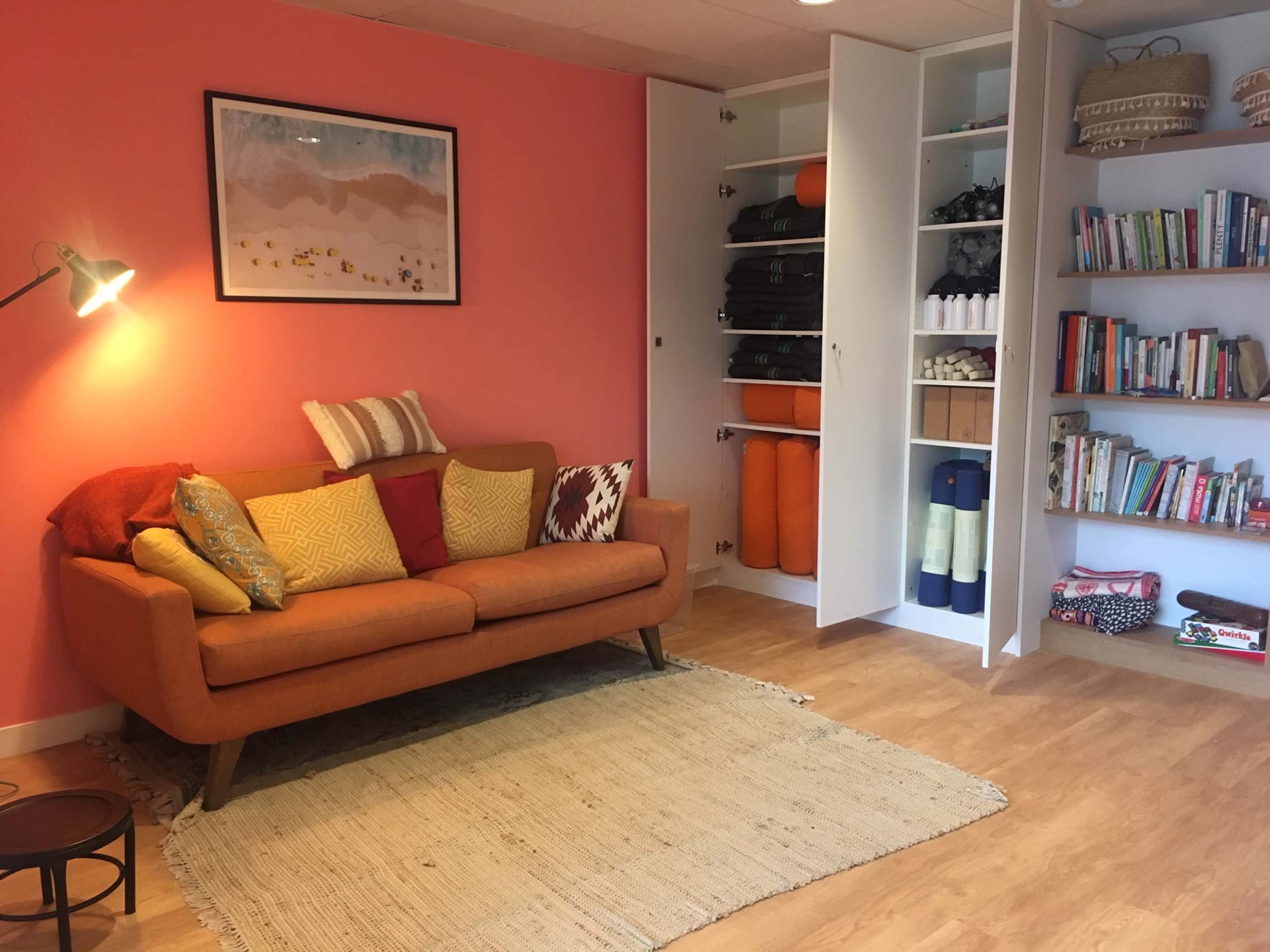 A bright living space features an orange sofa adorned with colorful pillows, a small side table, and a bookshelf filled with various books, alongside a storage cabinet.