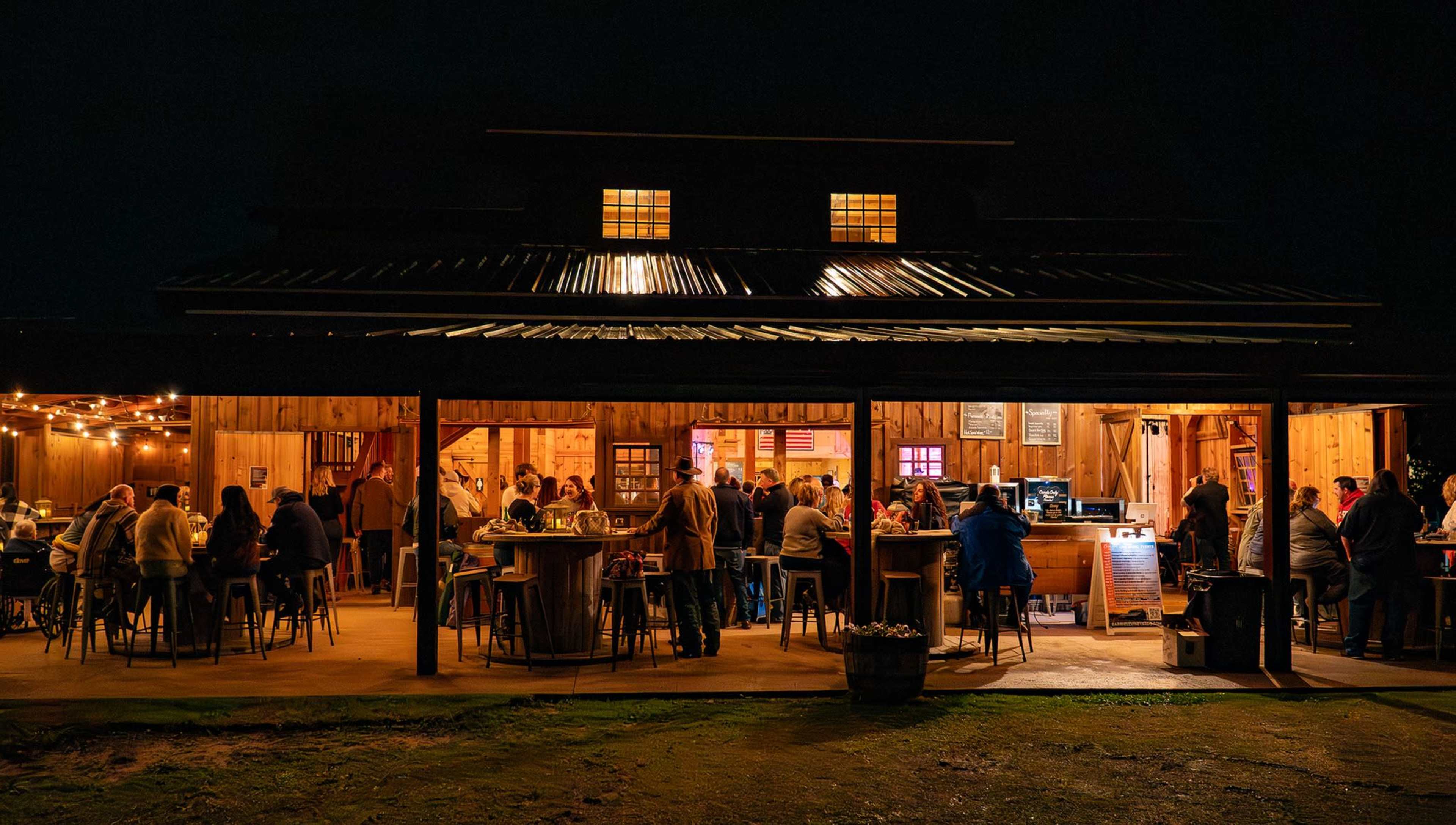 A lively crowd gathers at a rustic outdoor bar with a warm, inviting atmosphere illuminated by string lights and the glow from the interior.