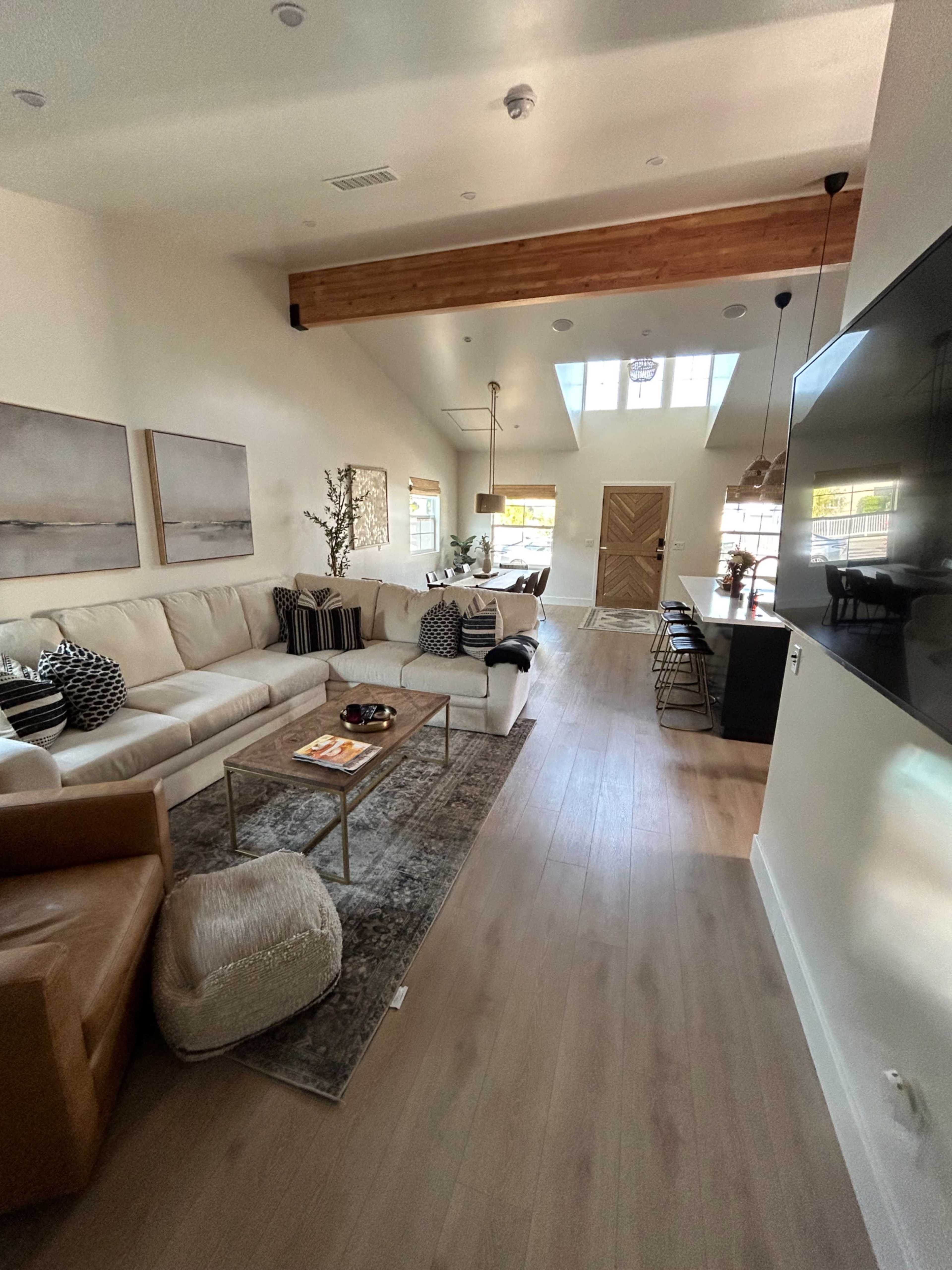 A modern living room with light-colored walls, a large sectional sofa, a coffee table, and a dining area in the background, featuring a high ceiling with wooden beams.